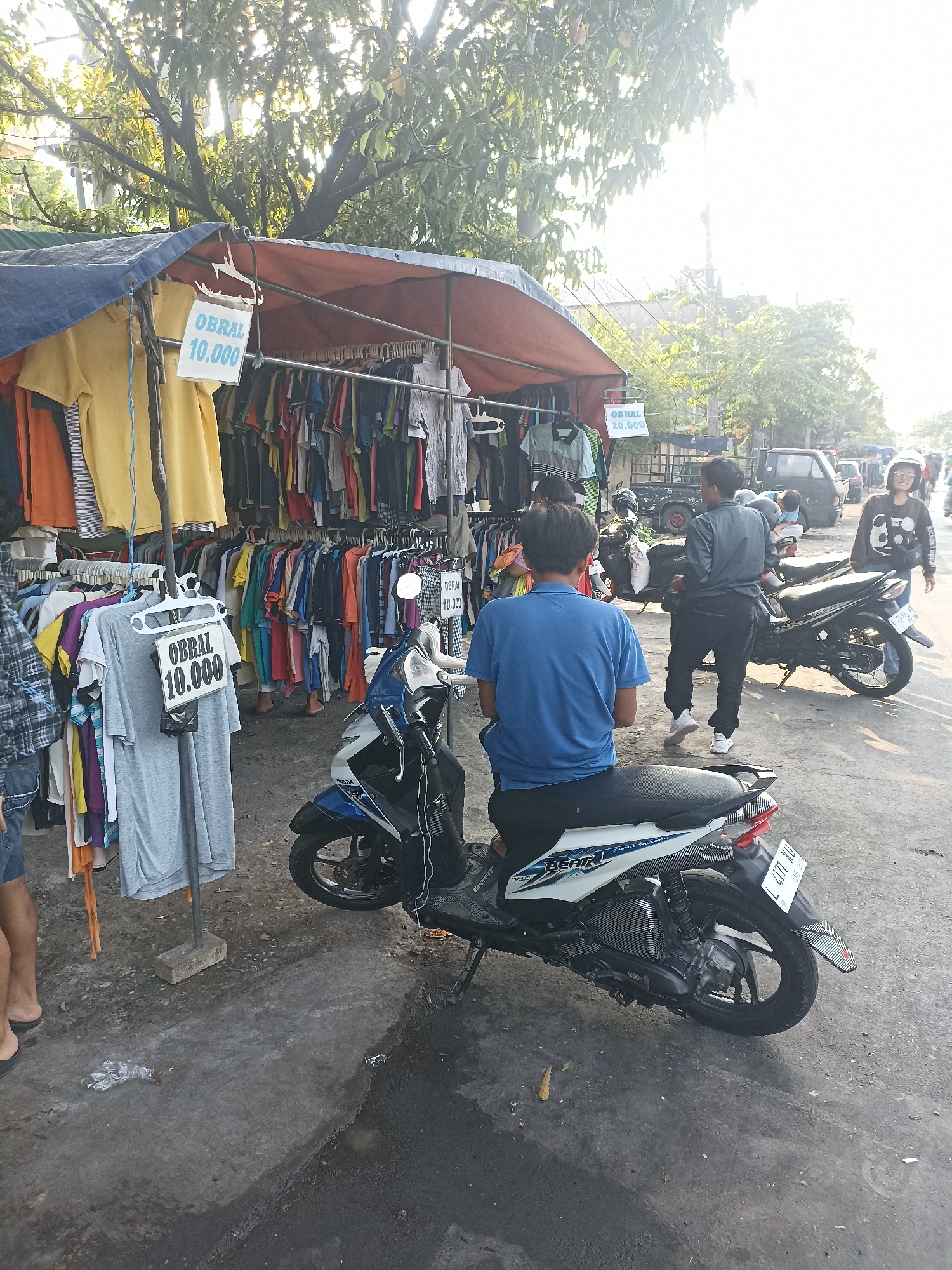 In Surabaya, Indonesia, an outdoor clothing stall is set up under a tarpaulin shelter, displaying an assortment of t-shirts and shirts on racks. Two price signs, marked "OBRAL 10.000," are visible, indicating a sale. A man in a blue t-shirt is seated on a white motorcycle parked in front of the stall. To the right, another man in a grey jacket stands near a black motorcycle, and further in the background, a woman in a black hoodie is walking. The scene appears to be a street market, with other vehicles and structures visible in the distance. The ground is paved and somewhat damp.