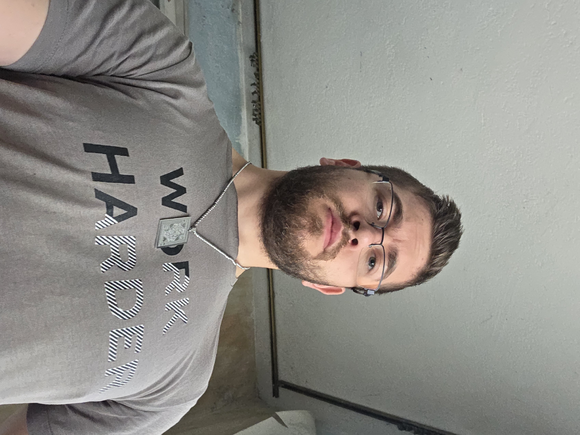 A fair-skinned male with a beard and glasses is depicted in an indoor setting. He is wearing a grey t-shirt with the phrase "Work Harder" printed on it and a necklace with a pendant. The background appears to be a simple wall. No specific details indicate the location is Yagodovo, Bulgaria.