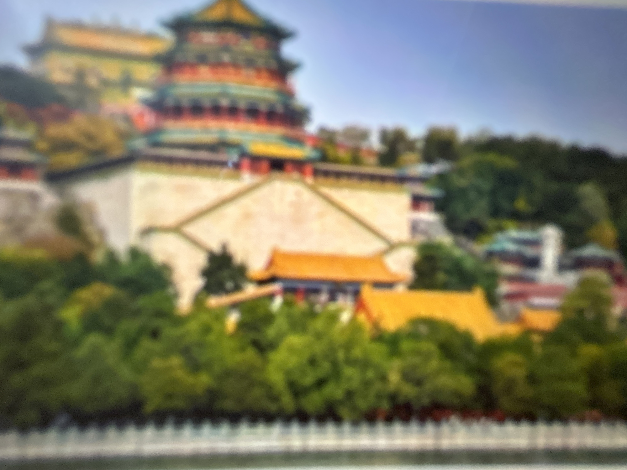 The image depicts a blurred, vertical view of a multi-tiered building with a vibrant, colorful roof, likely a pagoda or temple. The building is surrounded by lush green foliage, suggesting a park or garden setting. The sky appears clear and blue. The specific location in Gelendzhik, Russia, is not discernible from the provided image. There are no people or discernible actions visible in the scene.