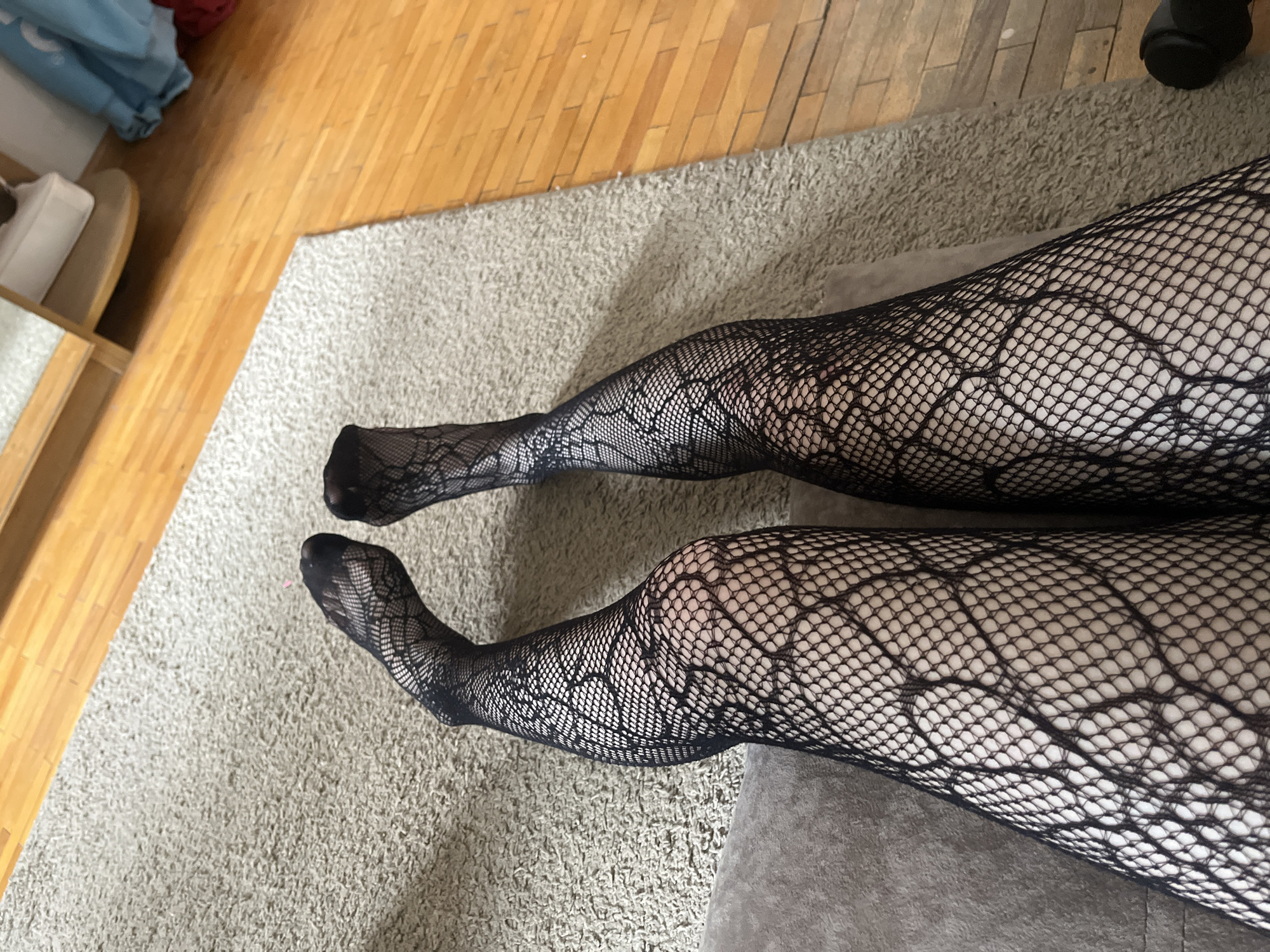 An overhead shot presents a pair of legs adorned in black fishnet stockings, resting in an indoor setting. The stockings feature a distinctive design, combining a fine fishnet mesh with a larger, elaborate lace-like pattern reminiscent of a floral or organic web. The person's skin is visible through the open weave of the hosiery. The legs appear relaxed, positioned on a soft, light grey shag carpet that covers most of the visible floor area. A section of a dark grey upholstered surface, likely a cushion from a couch or chair, is visible beneath the legs.
To the left and upper part of the frame, a smooth, light brown wooden floor is exposed, indicating a transition from the carpeted area. In the far background on the left, a white object, possibly a shoe, rests on a wooden shelf, with some light blue fabric partially visible nearby. The scene suggests a casual, domestic environment, such as a living room or bedroom. No specific activity is occurring beyond resting. The lighting is soft and ambient, characteristic of indoor conditions, and no time of day or external weather can be determined. There is no visible text. The location context provided for this image is Murino, Russia.