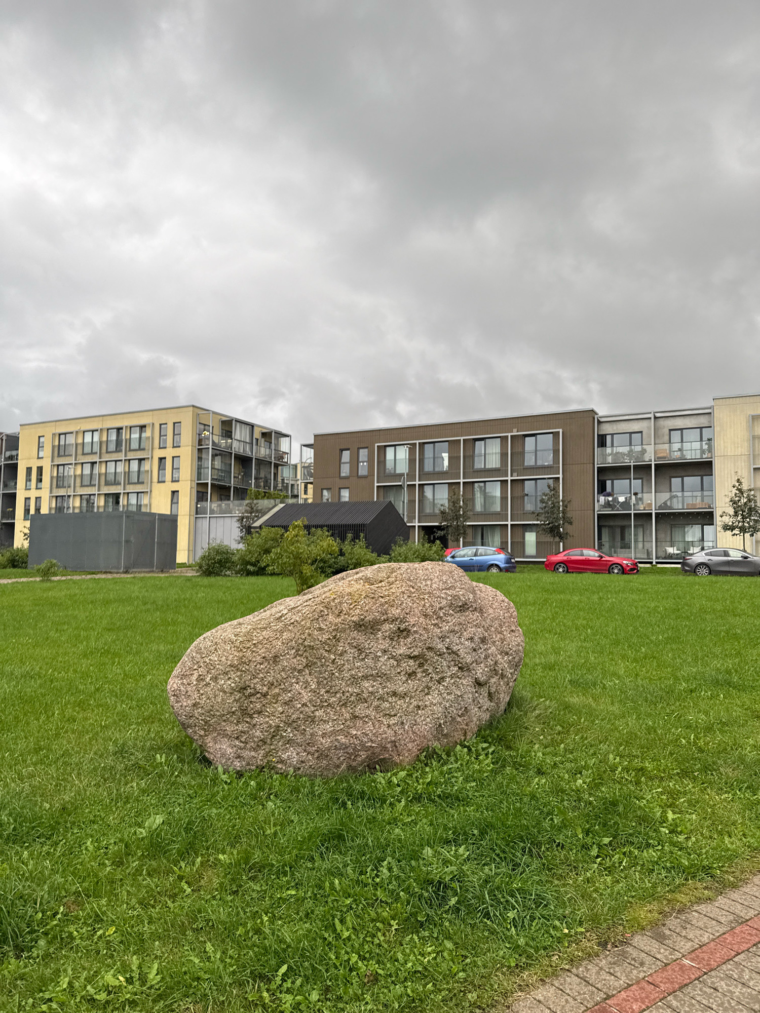 In Tallinn, Estonia, an outdoor scene is captured. The foreground is dominated by a large, rough-textured boulder situated on a lush green lawn. The grass is well-maintained, with some scattered weeds. To the left and behind the boulder, a grey construction fence is visible. In the midground, several modern apartment buildings line the horizon. The building on the far left is yellow and features balconies and numerous windows. Adjacent to it is a darker, brown building with a similar architectural style, also equipped with balconies. Further to the right, another section of an apartment building with light-colored facades and balconies is visible. Several cars are parked in front of the buildings: a blue hatchback, a red sedan, and a grey sedan. The background is an overcast sky with a diffuse light, indicating a cloudy day.
