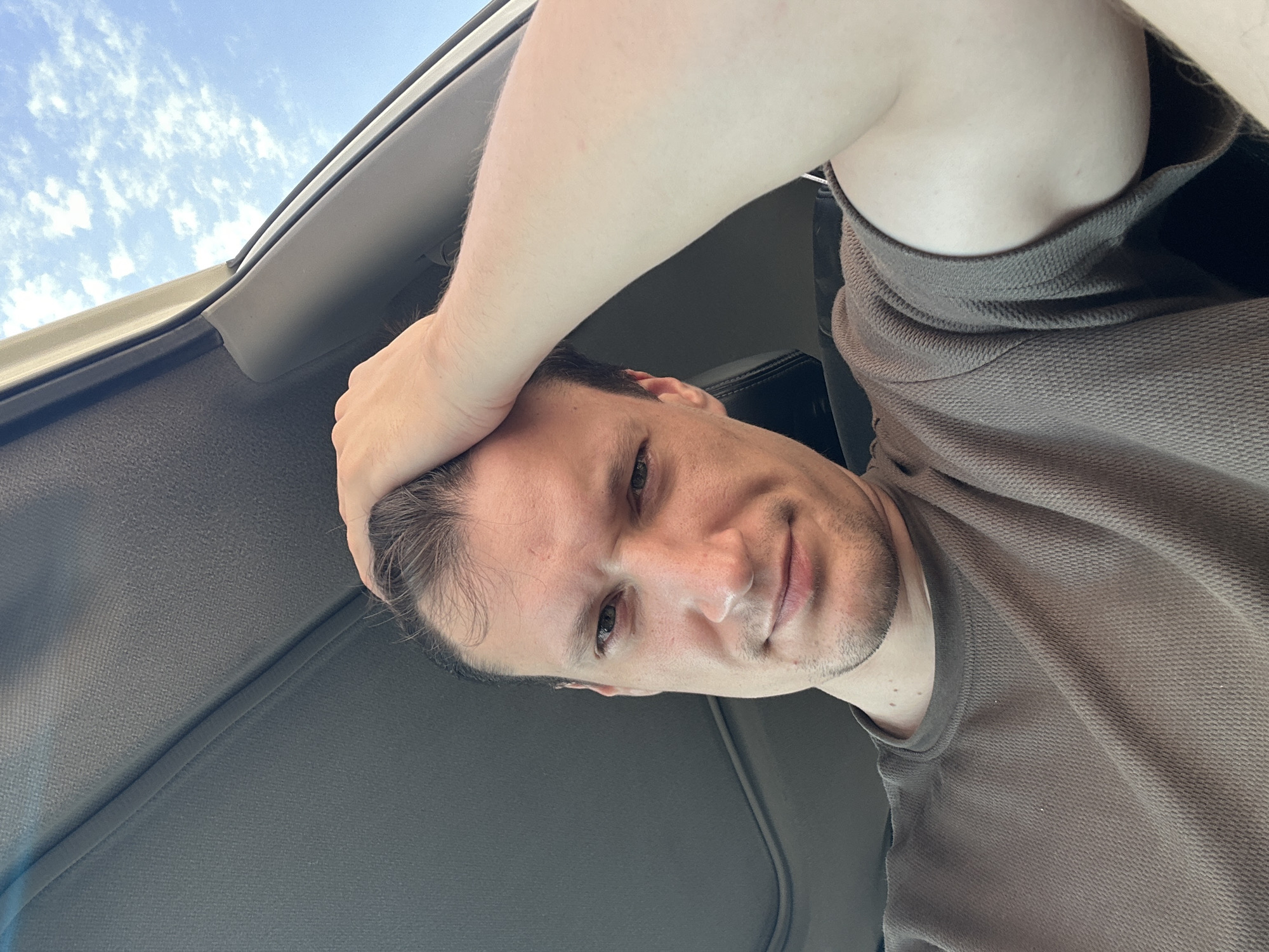 A man is sitting in a car with his hand on his head. He is wearing a brown shirt. The car appears to be a sedan, with the sunroof open to reveal a blue sky with white clouds. The image is taken from a first-person perspective, looking up at the man and the sky. There is no indication of Shatava, Ukraine in the image.