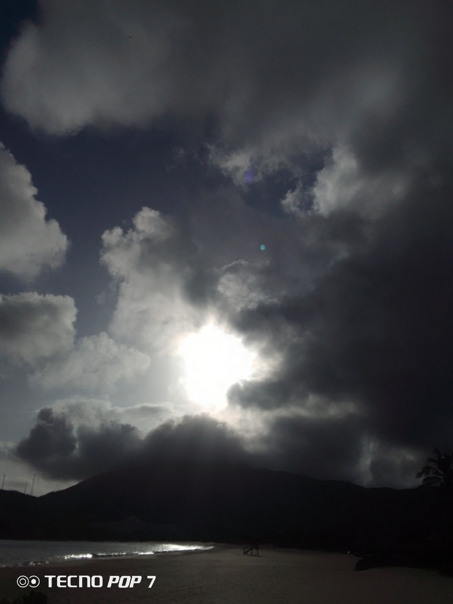 The photograph depicts a beach scene at Pedro González, Venezuela. The image is dominated by a sky filled with dramatic, heavy clouds, partially obscuring the sun. The sun's rays break through the cloud cover, creating a bright spot. Below the sky, a dark silhouette of a mountainous or hilly landscape is visible. In the foreground, there is a sandy beach and the edge of the water with some glimmers of light. The overall atmosphere is dark and moody. The camera used, a TECNO POP 7, is written in the bottom corner.