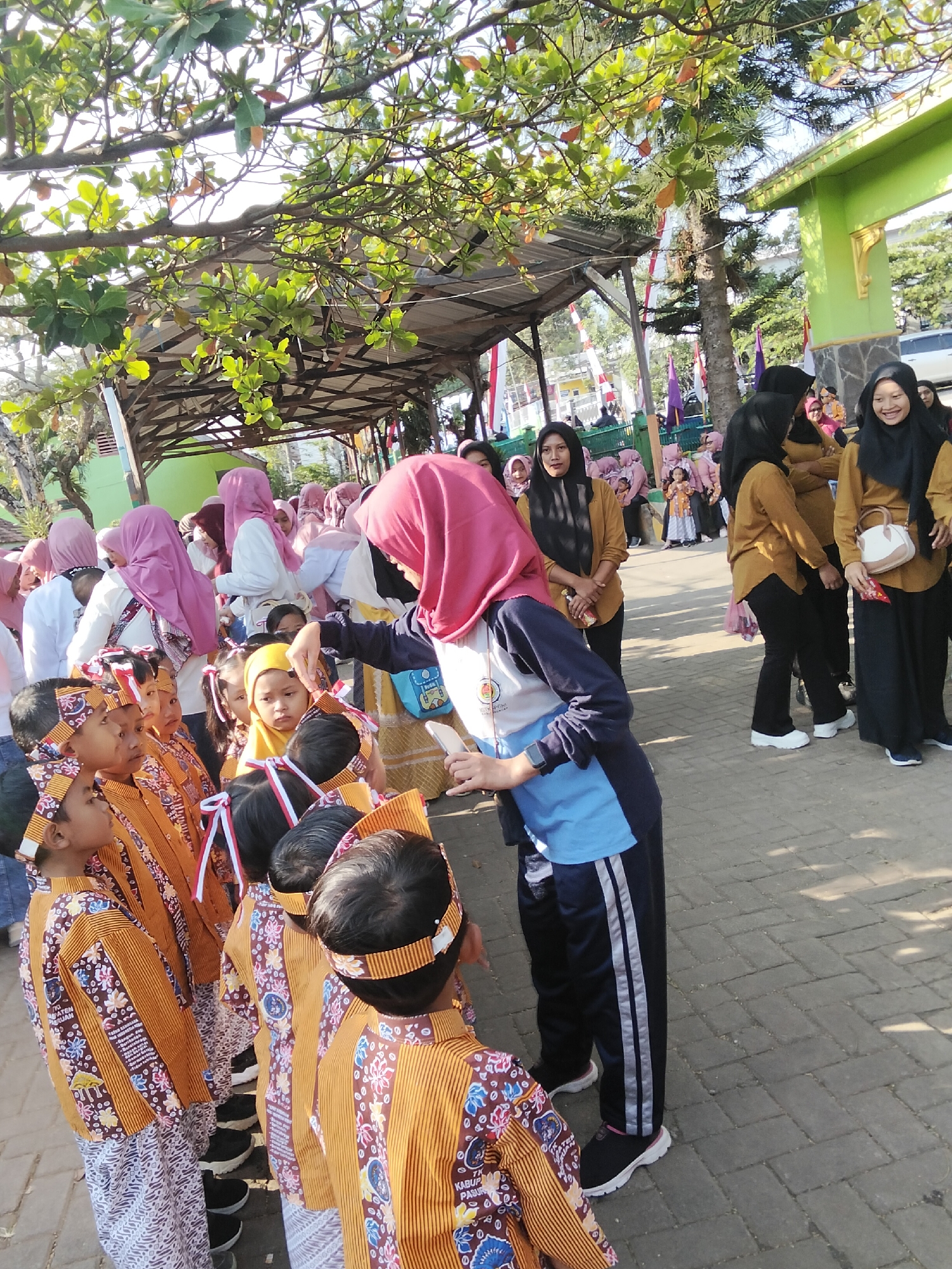 In Puspo, Indonesia, a group of children in traditional batik attire stand in a line outdoors, facing a woman in a pink headscarf and blue and white sportswear. The children wear headbands with red and white ribbons. The woman appears to be interacting with one of the children, possibly handing something to them. Other individuals, including adults in hijabs and traditional clothing, are visible in the background, along with trees and a green building. The scene suggests an outdoor event or gathering.