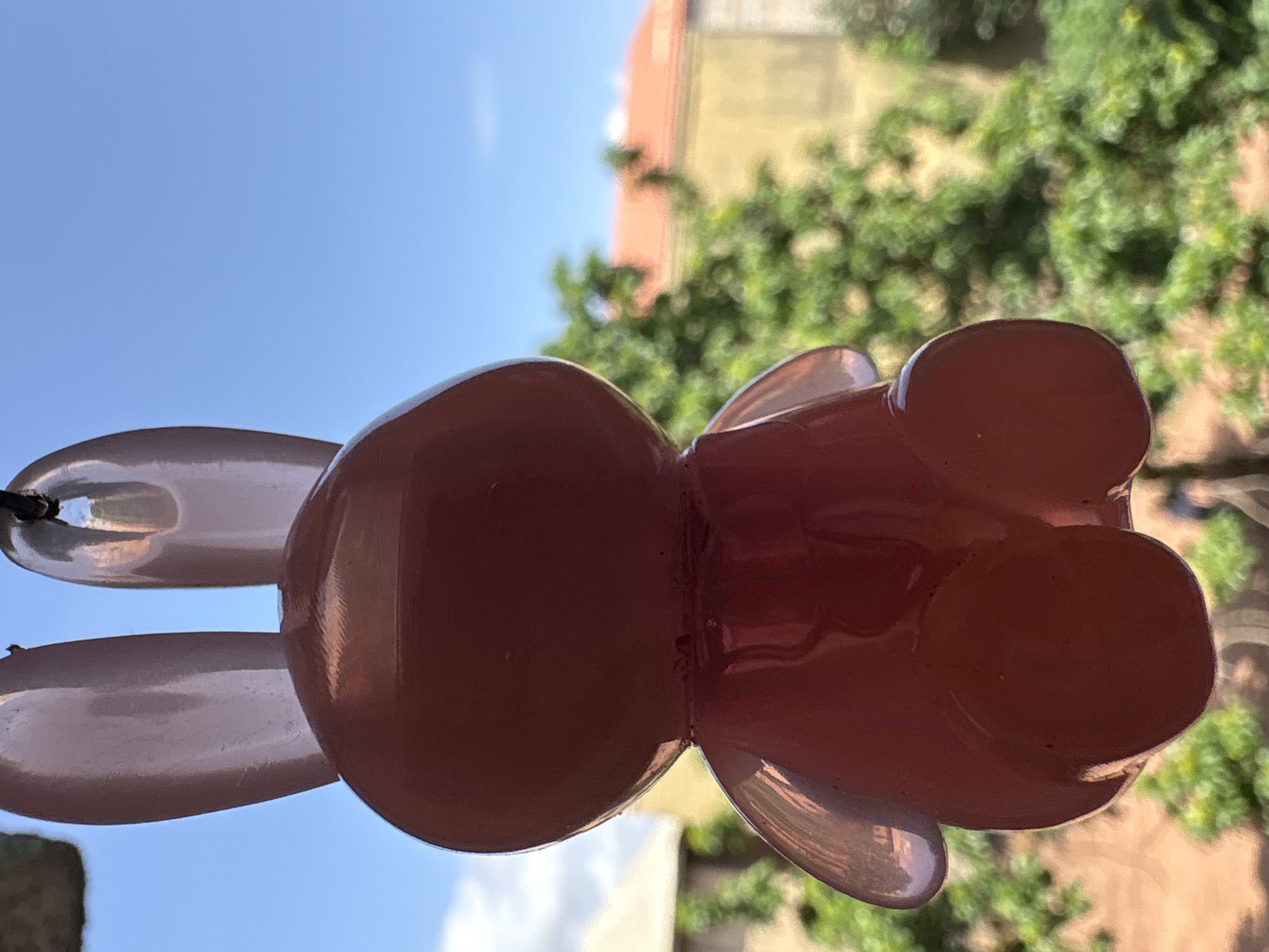 A pink, translucent resin figurine of a rabbit is hanging outdoors, possibly as a decoration or wind chime. The figurine is positioned in the foreground, obscuring the view of the background. The background consists of a bright blue sky with some clouds, lush green trees, and what appear to be buildings with orange and yellow roofs. The image was taken in Onitsha, Nigeria.