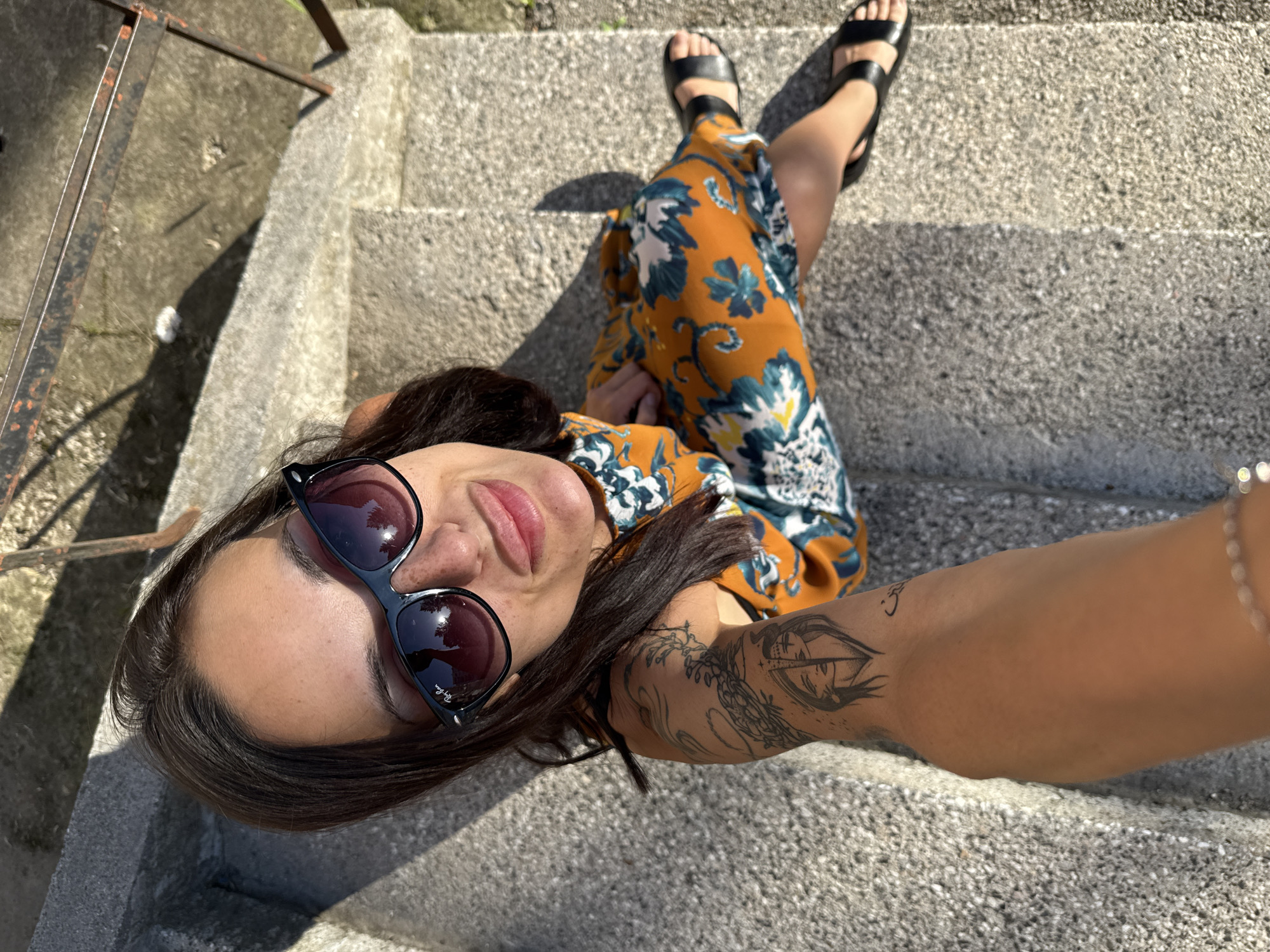 A woman is sitting on concrete stairs, taking a selfie from an overhead angle. She is wearing sunglasses and a floral dress, and has tattoos on her arm. She is wearing black sandals. The location is an outdoor setting, specifically concrete stairs that appear to be in Pleven, Bulgaria.