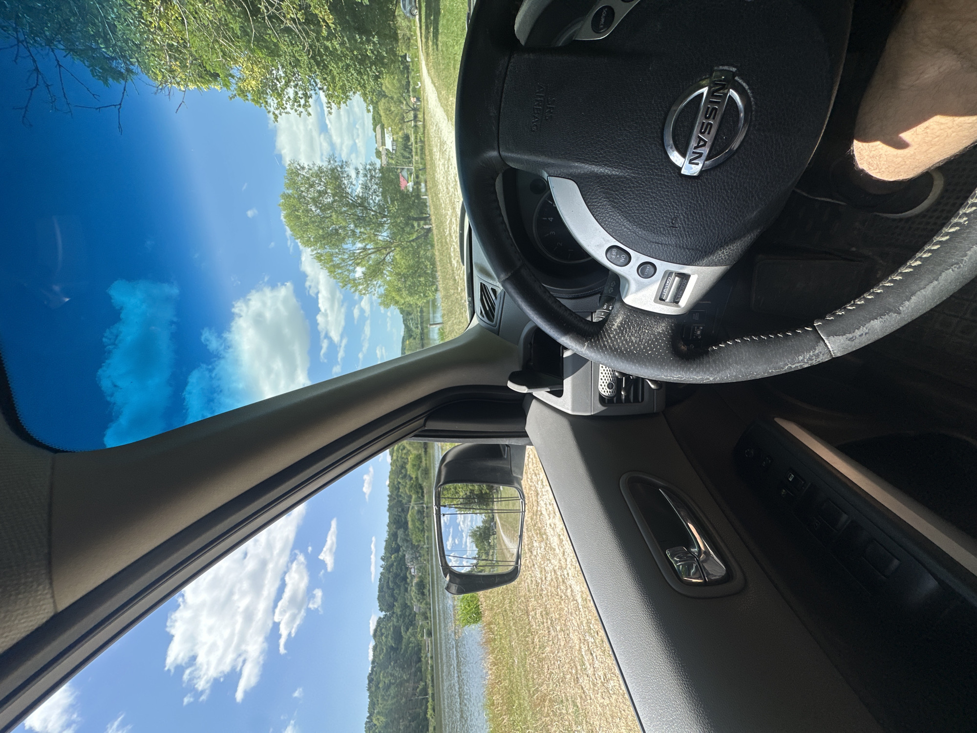 The image, taken from inside a Nissan vehicle, shows a view through the driver's side window and windshield. Outside, a body of water, possibly a river or lake, is visible, bordered by grassy banks and a treeline. The sky is blue with scattered white clouds. The interior of the car includes the steering wheel, dashboard, door panel with window controls, and a portion of the driver's leg. A side-view mirror reflects the scenery outside, including a path and trees. The image is set in Makiv, Ukraine.