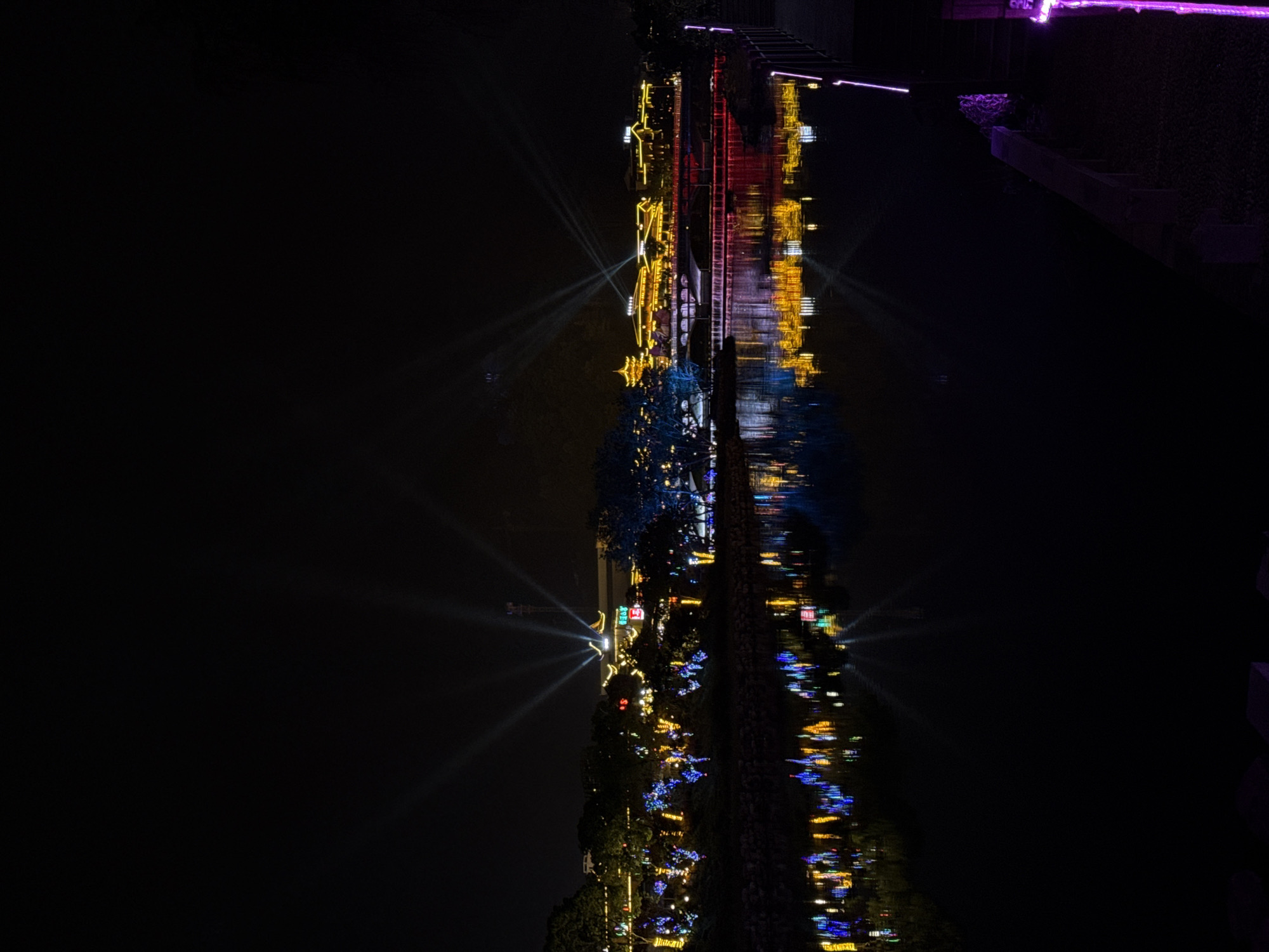 A nighttime shot depicts a body of water reflecting the illuminated architecture and landscaping of Wulingyuan, China. On the right, a wooden walkway is illuminated by purple lights. Towards the middle, buildings with yellow, red, and white lights are reflected in the water. A bright light with rays emanating upward stands out among the lights. The reflections appear somewhat distorted, indicating movement in the water. A large dark area occupies the left portion of the image.