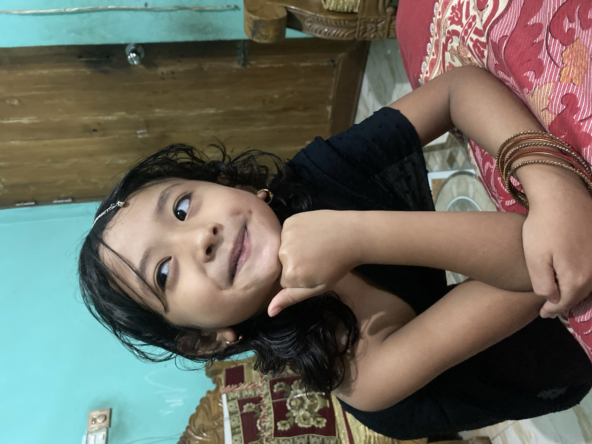 A young girl with long, dark, wavy hair, wearing a black garment and bangles on her right wrist, is leaning on her right arm, propping her chin on her fist. She is looking up and to her right with a slight smile. She has a small ornament on her forehead. She is positioned in what appears to be a bedroom. The walls are painted a light blue-green color. To the left, a carved wooden headboard of a bed is visible, with a maroon and gold patterned fabric. To the right, a wooden door with a silver doorknob is present. The girl is sitting on a bed with a red and white patterned bedspread. The overall setting suggests a domestic interior, likely in Dhaka, Bangladesh, given the context provided.