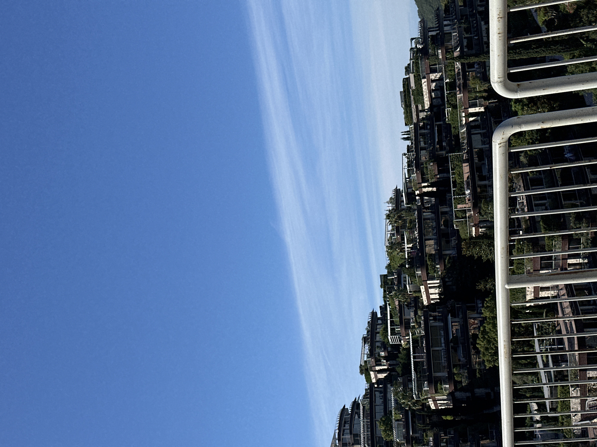 The image displays a terraced hillside residential complex with a modern architectural style, situated in Budva, Montenegro. The buildings are multi-level structures with balconies, glass windows, and lush greenery integrated into their design, creating a harmonious blend with the natural environment. The complex appears to be built into the slope of a hill, with each level cascading down. The foreground features a white metal railing, suggesting the vantage point is from a balcony or walkway. The sky above is a clear, vibrant blue with wispy white clouds. The overall scene conveys a sense of luxury and contemporary living within a scenic coastal landscape.