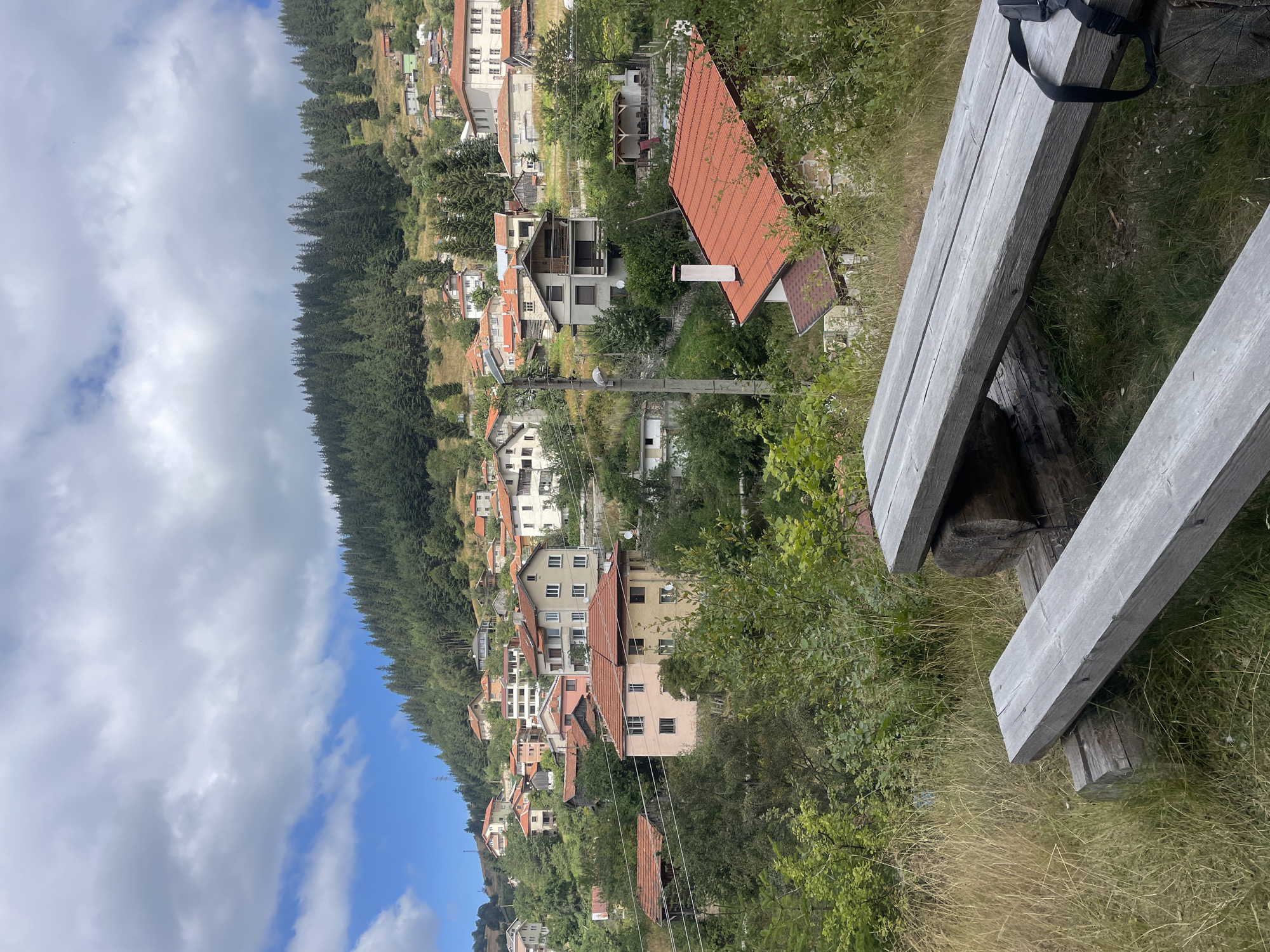 The image depicts a view of the village of Manastir, Bulgaria, nestled on a hillside covered in dense evergreen forest. In the foreground, a rustic wooden picnic table and bench are situated on a grassy knoll. The table is made of rough-hewn logs and planks. Beyond the picnic area, the village unfolds across the undulating terrain, with numerous houses featuring red-tiled roofs. The architecture appears to be a mix of traditional and more modern styles, with varying colors and sizes of buildings. Some houses are visible nestled amongst trees, while others are more exposed on the hillside. The sky above is partly cloudy, with patches of blue sky visible between dramatic white and gray clouds. Power lines are also visible stretching across the scene. The overall impression is one of a peaceful, rural mountain village.