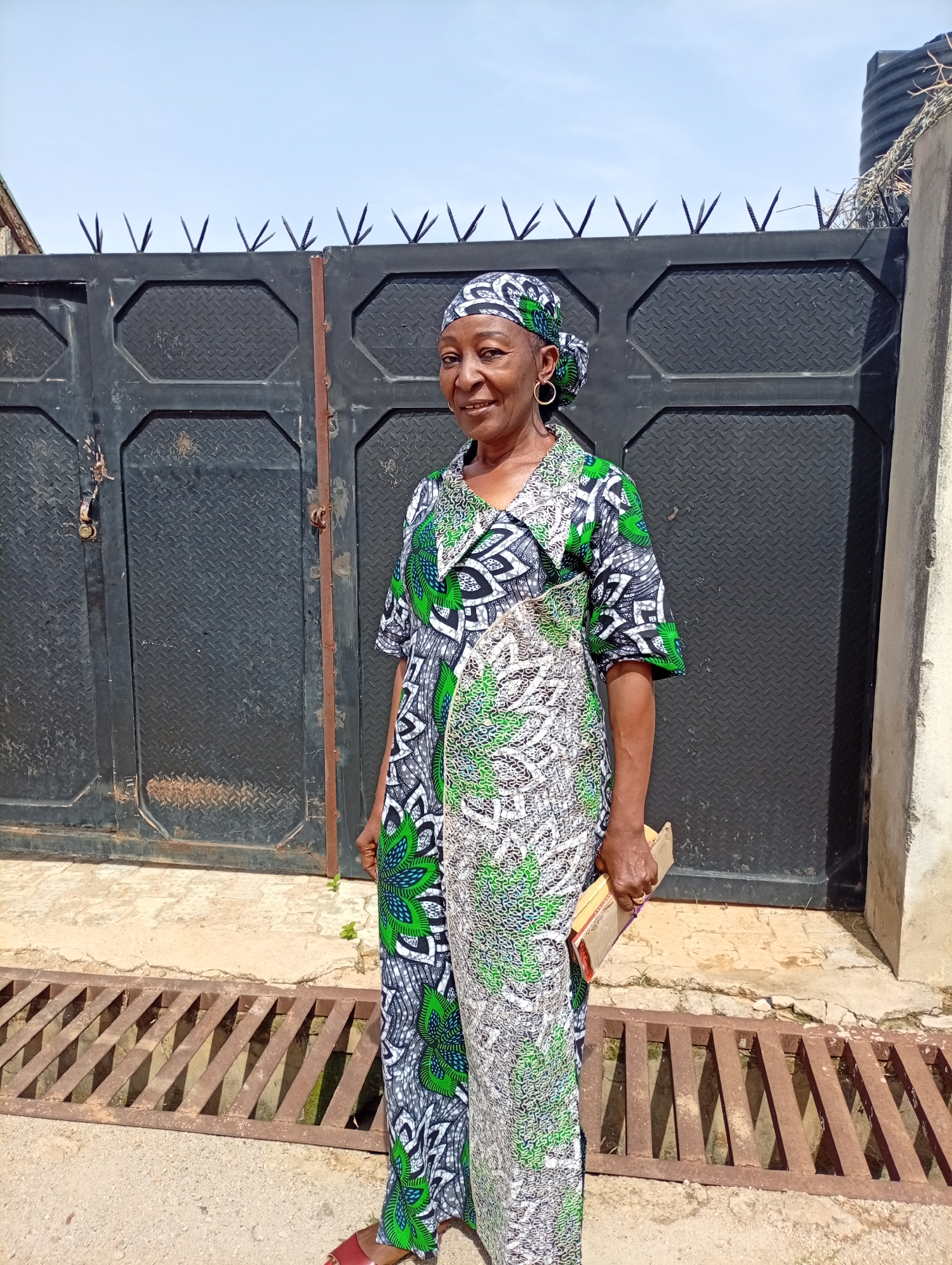 An adult female stands in front of a black metal gate. The gate has pointed, triangular spikes along its top edge. The woman wears a patterned green, gray, and white dress and a matching headscarf. She holds a book in her right hand. The location appears to be an outdoor area in a city; a metal drain grate is visible on the ground in front of her. The city and region are Unknown, Unknown.