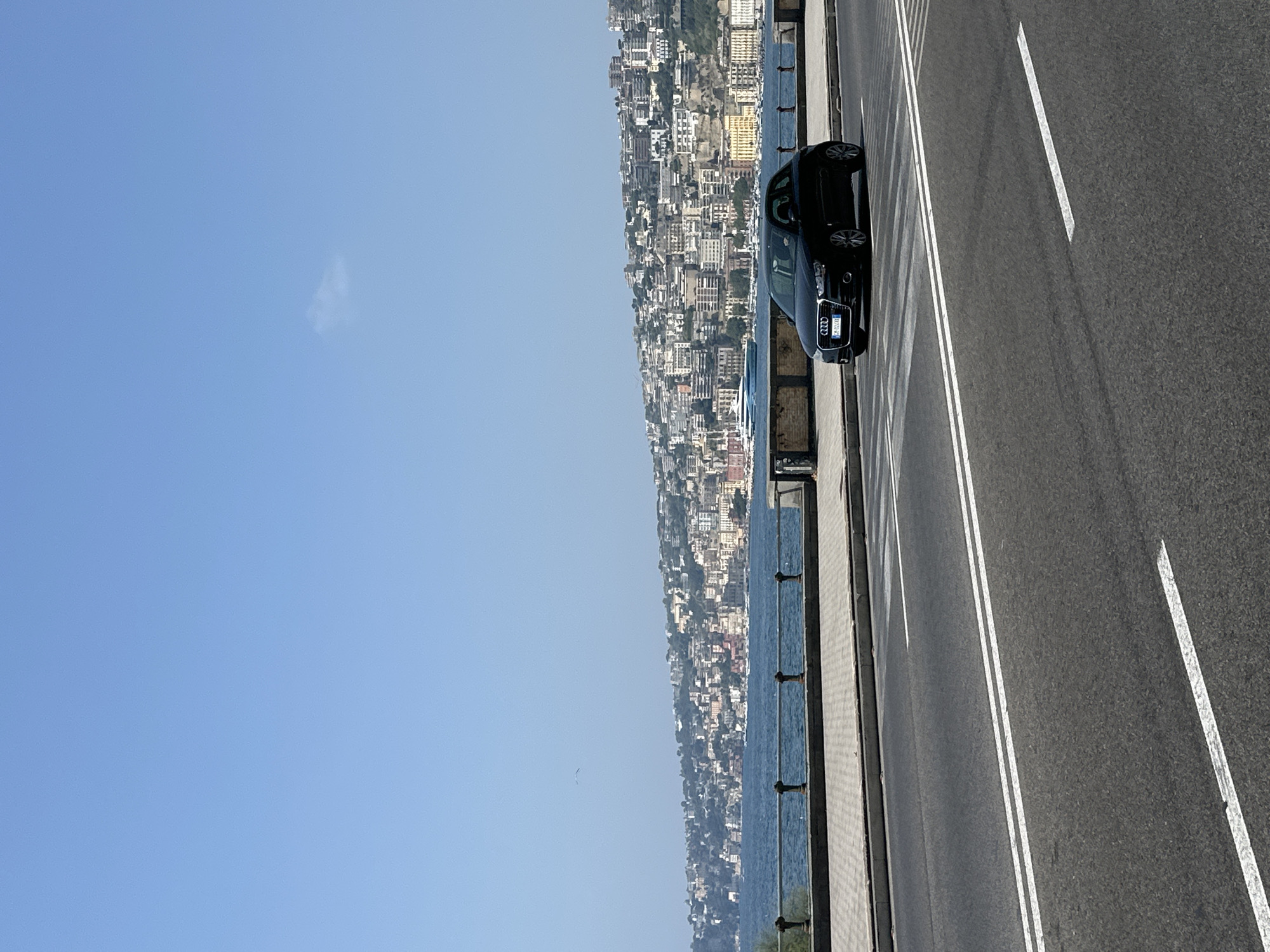 The image depicts a scenic view of the city of Naples, Italy, with the Bay of Naples in the background. The foreground features a two-lane asphalt road with white lane markings. A black Audi A1 hatchback is parked or driving on the right lane of the road.
In the middle ground, a promenade with a stone pavement and a decorative metal railing runs parallel to the bay. Beyond the promenade, the deep blue waters of the Bay of Naples are visible, with several boats and a large white ferry or cruise ship in the distance.
The background is dominated by a densely populated hillside covered with buildings of varying architectural styles and colors, characteristic of Naples. The buildings are packed closely together, creating a picturesque urban landscape that slopes down towards the sea. The sky is clear and bright blue, with a few wispy clouds. The overall impression is one of a sunny day in a vibrant coastal city.