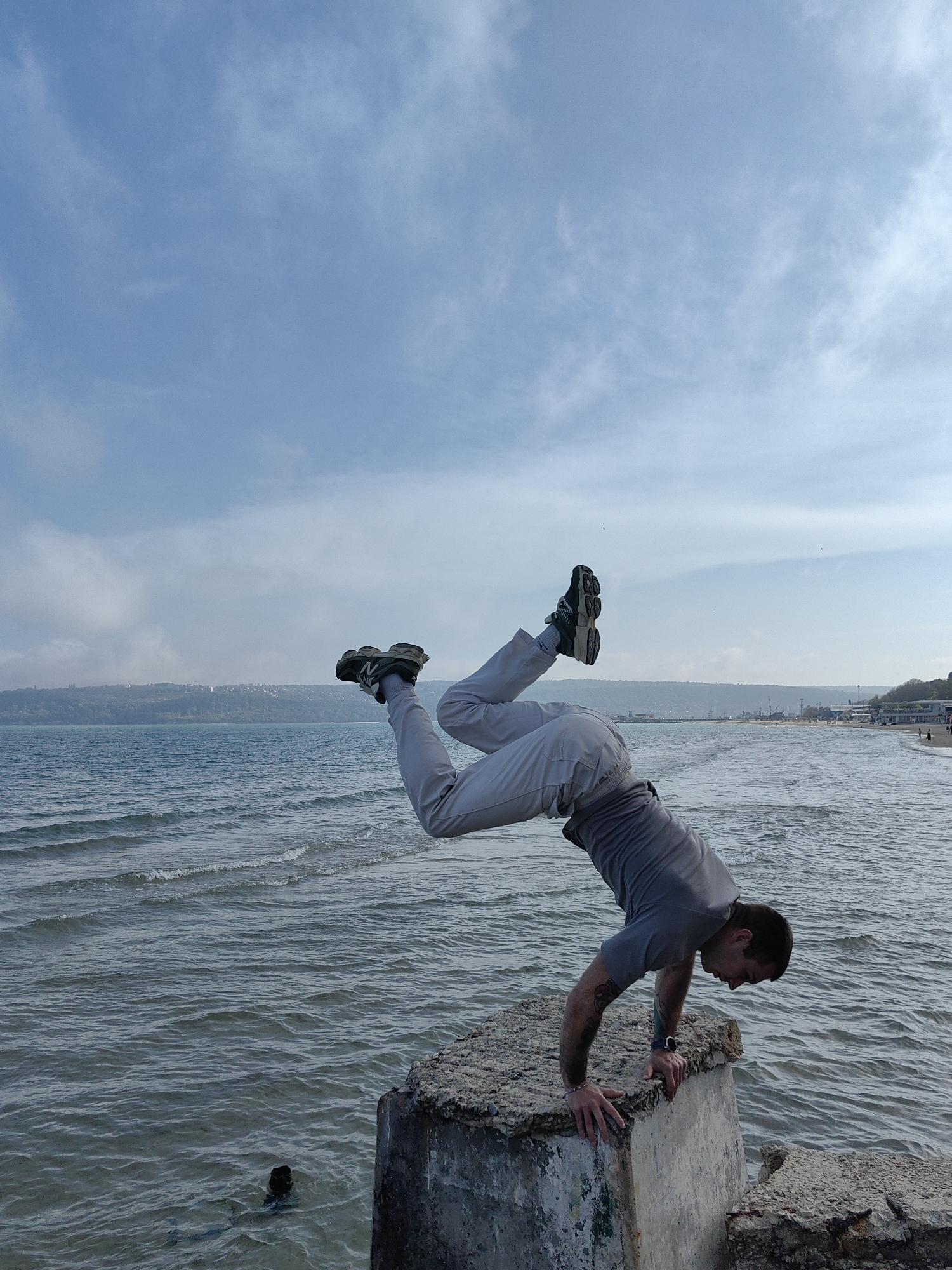 A man is performing a handstand on a concrete structure near the sea in Varna, Bulgaria. He is wearing a grey t-shirt, light grey pants, and dark-colored sneakers with white socks. His body is positioned horizontally with his legs slightly bent and lifted. He has a tattoo on his left arm and a watch on his left wrist. The concrete structure is situated at the edge of the water. The sea is slightly choppy, with small waves, and the background reveals a coastline with some buildings and trees. The sky is blue with scattered clouds. There is a dark object in the water near the concrete structure.