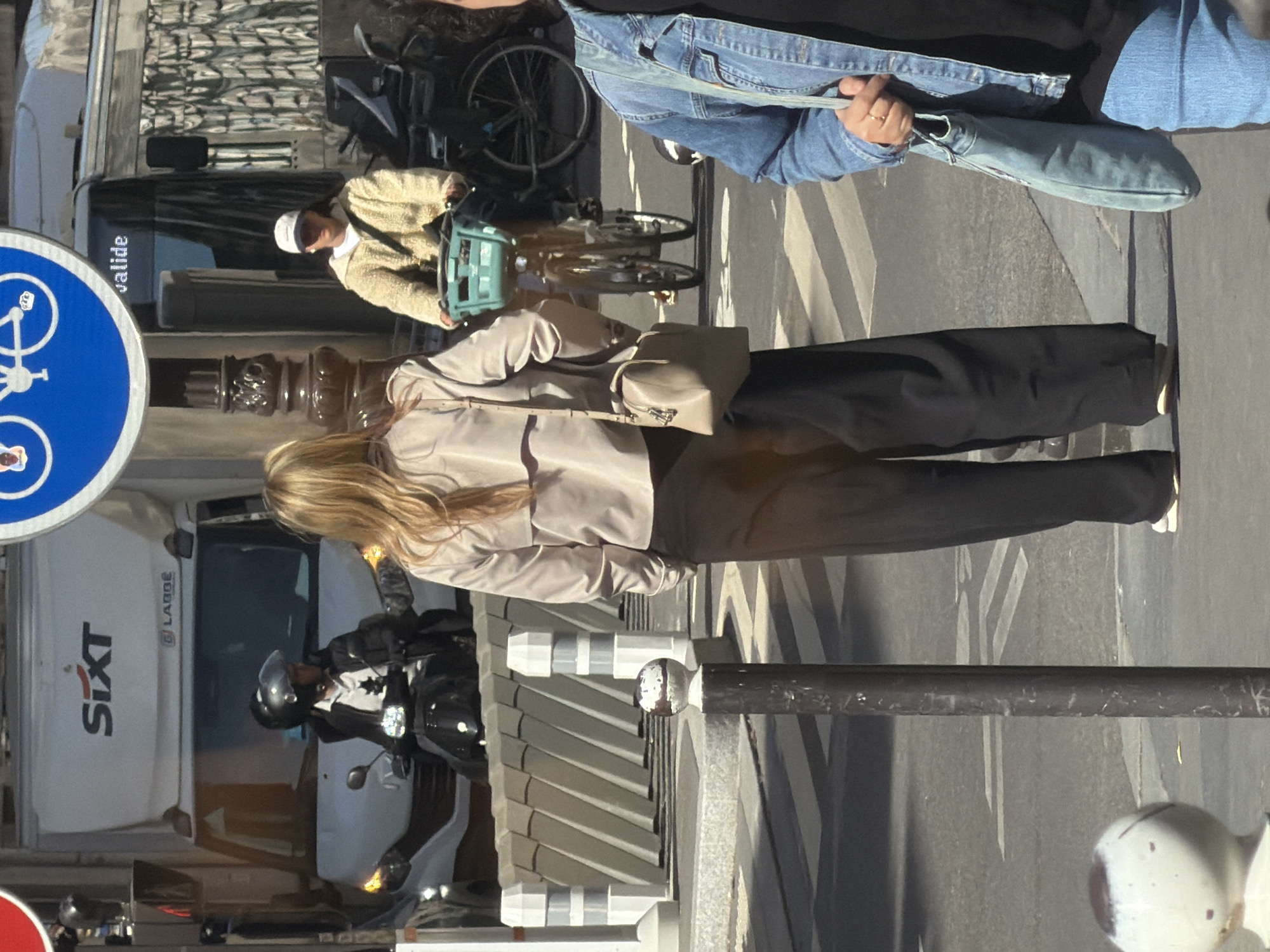 A street scene in Paris, France, shows a person with blonde hair, wearing a beige jacket and black pants, walking on the sidewalk, with a light beige crossbody bag on their shoulder. Next to them is a black metal pole and a partially visible white structure with a pitched roof. To the left, a white van marked "SIXT" is partially visible, with a man on a motorcycle wearing a helmet standing beside it. In the background, a cyclist in a light-colored jacket and white cap rides past a dark-colored car. A person wearing a denim jacket stands on the sidewalk. A blue bicycle sign is prominently displayed in the foreground, and the street is marked with white lines.