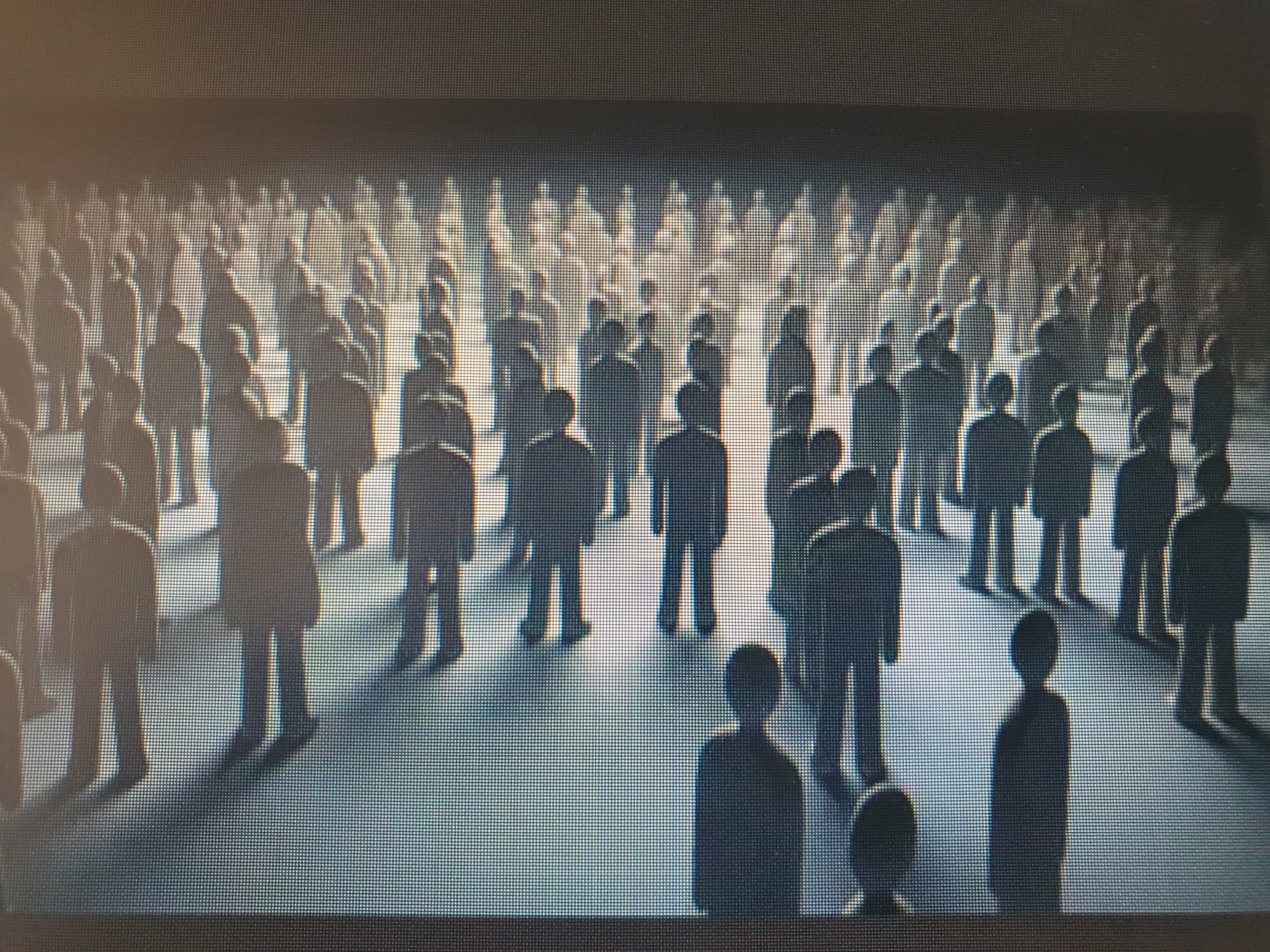 The image shows a dimly lit scene with a large crowd of silhouetted human figures. The figures appear to be standing and are uniformly depicted as dark shapes against a lighter background, suggesting they are not individually detailed. The lighting creates long shadows stretching across the ground, indicating a low light source, possibly from the side or behind the viewer. The environment appears to be a flat, open space, with the figures arranged in rows and clusters, implying a gathering or assembly. The overall impression is one of uniformity and perhaps anonymity due to the lack of individual features. There is no specific indication of the location being the city of Tashkent, Uzbekistan.