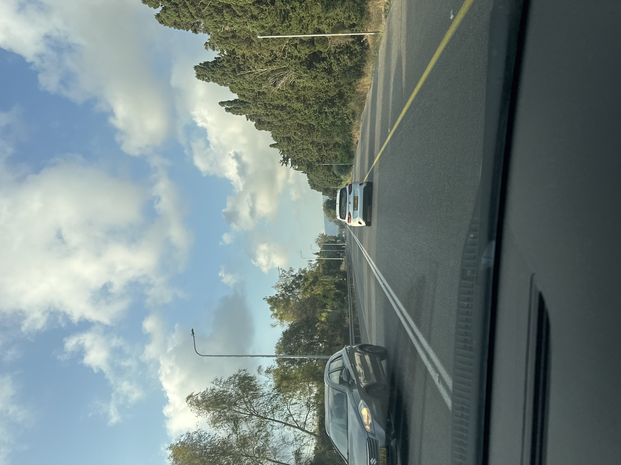 The image captures a daytime scene on a multi-lane highway in Nahariyya, Israel. The perspective is from inside a vehicle, looking forward. On the left side of the road, a dark-colored SUV is visible in the foreground, driving in the same direction. Further ahead, a white hatchback is also traveling on the highway. The road is bordered by a line of tall, dense cypress trees on the right and a row of deciduous trees on the left. The sky above is a mix of blue with scattered white clouds, suggesting partly cloudy conditions. Streetlights are present along the side of the road at regular intervals. The asphalt road has clear lane markings, including a yellow line dividing opposing traffic and white lines separating lanes in the same direction. The overall environment appears to be a rural or semi-rural section of highway.