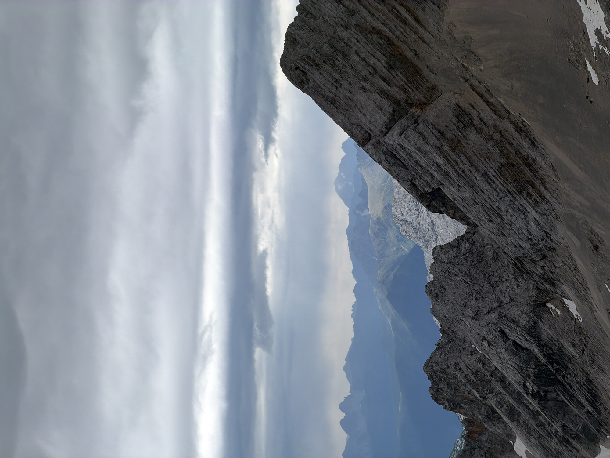 This is a landscape photograph taken from a high vantage point in the Bavarian Alps, likely on the Zugspitze, the highest peak in Germany, near Garmisch-Partenkirchen. The foreground is dominated by the rugged, gray, and rocky slopes of a mountain range, with patches of snow visible in the crevices and on some of the lower slopes. The texture of the rock is highly detailed, showing striations and rough surfaces.
In the middle ground, successive layers of blue-tinged mountain ranges recede into the distance. These ranges appear more rounded and softened by atmospheric perspective compared to the sharp, detailed foreground. Some of these distant mountains have green slopes, suggesting lower altitudes or valleys.
The sky is overcast with heavy, dark gray clouds that obscure the sun. There are breaks in the clouds through which shafts of light appear to be filtering down, creating a dramatic and somber atmosphere. The lighting is diffused, casting a general gloom over the scene, though the light beams add some dynamism. There are no people or man-made objects visible in the image. The overall impression is one of wild, imposing, and somewhat bleak natural beauty.