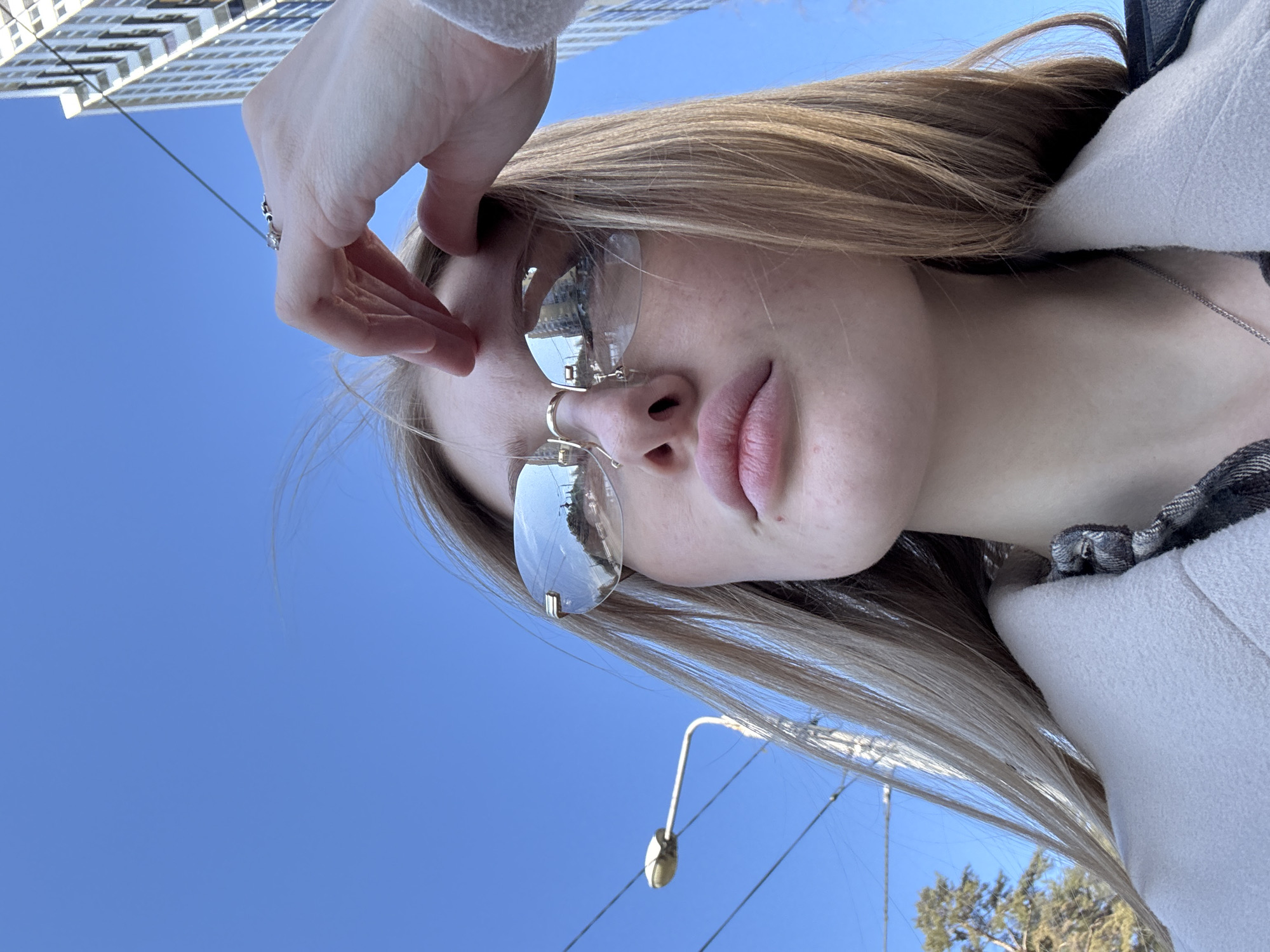 A low-angle, close-up shot captures a young individual, likely female, in an urban environment in Perm, Russia, under a clear blue sky. The subject possesses fair skin and long, straight, light-colored hair, with some strands appearing wind-blown. She wears rimless, reflective sunglasses featuring gold-toned bridge and temple details, which show reflections of urban structures and roads. Her lips are full. She is dressed in a light-colored, possibly off-white, coat worn over a dark, patterned garment, and a thin silver-colored chain necklace is visible. Her right hand is raised to her forehead, fingers slightly splayed, displaying a ring with a prominent stone and a patterned band on the ring finger. The background features a street lamp pole and overhead utility wires to the left, accompanied by bare tree branches and a segment of a pine tree. To the right, a tall, modern building with a gridded facade rises against the sky.