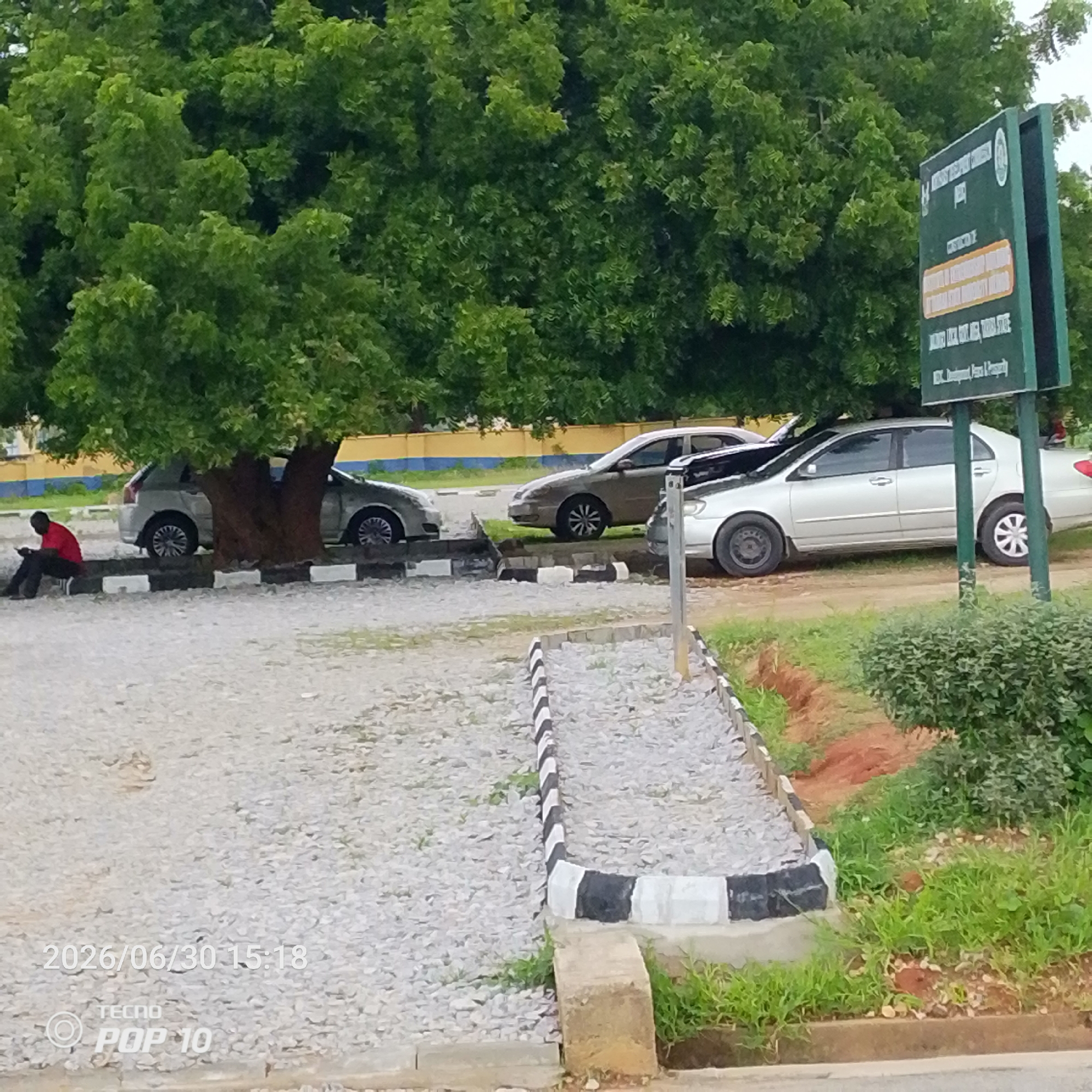 The image depicts an outdoor scene in Jalingo, Nigeria. In the foreground, a person wearing a red shirt and black pants is seated on a low wall, looking down at a mobile device. Behind the person, several cars are parked under the shade of a large tree. One car is partially visible to the left, and two others are more clearly seen to the right, one silver sedan and another light-colored sedan.
The ground is covered in light-colored gravel. Black and white striped concrete barriers line the parking area. In the background, there is a yellow and blue painted wall, suggesting the area might be adjacent to a building or institution. A green signpost with white lettering is visible on the right side of the frame, partially obscured by foliage. The sign has several lines of text, with the top line appearing to be a name or title, followed by smaller text below.
The foliage of the large tree is dense and green, indicating a warm climate. The sky is overcast, suggesting a cloudy or possibly rainy day. The lighting is diffused, typical of an overcast sky.
Visible text includes a timestamp in the bottom left corner: "2026/06/30 15:18" and "TECNO POP 10". The signpost also contains legible text, though the full content is not clear due to the resolution and angle.
The overall impression is a quiet, casual moment with people and vehicles in a shaded outdoor area. The presence of the signpost and the organized parking suggests a public or institutional setting.