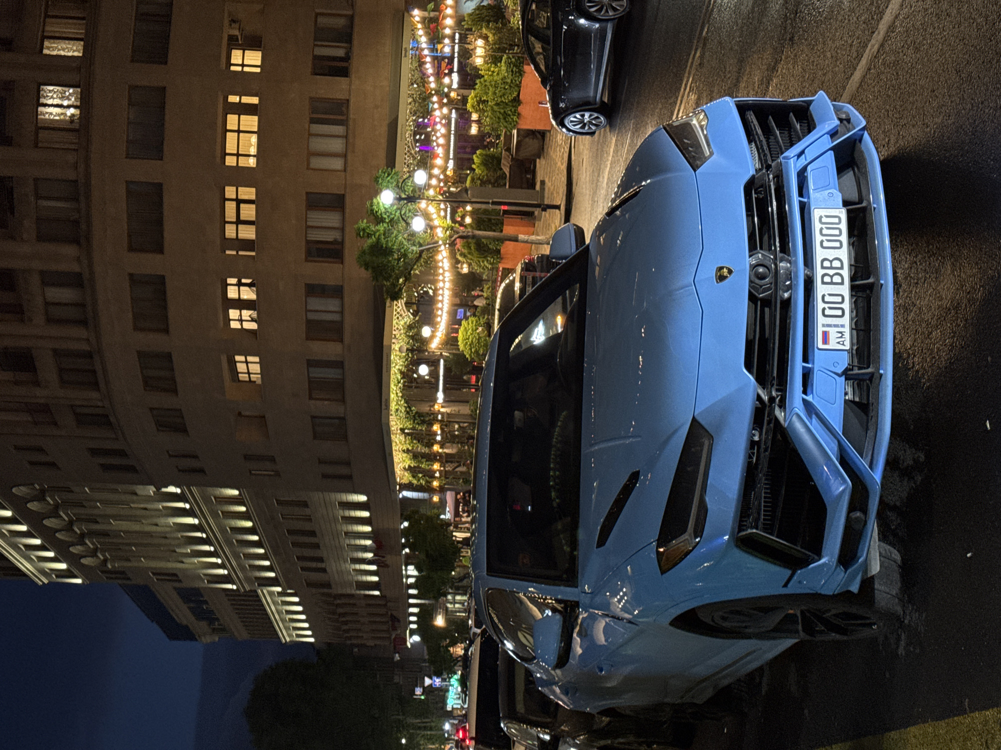 The image shows a light blue Lamborghini Urus parked on a street in Yerevan, Armenia at night. The car has a license plate with "00 BB 000" in Armenian script. The building behind the car has many windows lit up and illuminated architectural details. In the background, there's a cafe or outdoor dining area lit with string lights.
