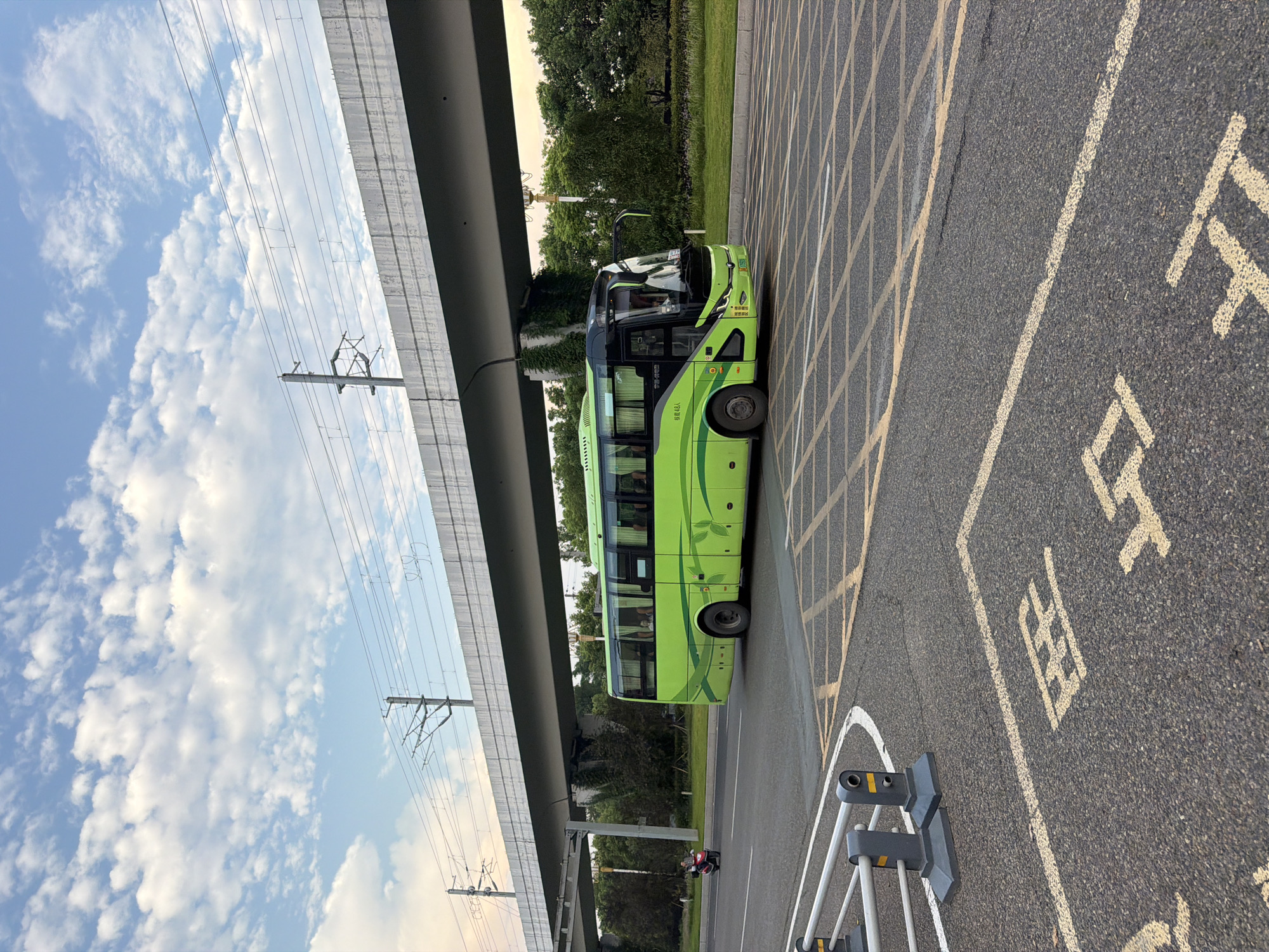 A bright green tour bus is driving on a road in Jiangjin, China. The bus is in the right lane of a two-lane road. There is a raised highway overpass with overhead power lines. The bus has tinted windows and a black roof. On the side of the bus are some leafy green patterns. In the distance, there are trees and green grass. On the road in the foreground, there are yellow and white painted lines and markings. There is also a motorcycle with a rider going in the opposite direction of the bus. The sky is blue with white clouds.