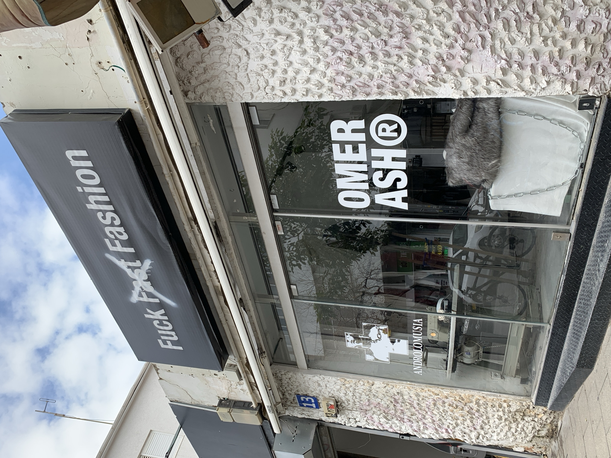 Here is a description of the image:
The image shows the storefront of a clothing shop in Tel Aviv, Israel. The awning above the store is black with white text reading "Fuck Fast Fashion" with an "X" painted over "Fast." The shop's name, "OMER ASH®", is displayed prominently on the glass storefront. The address number "13" is visible next to a sign with the name "ANDROLOMUSIA." Inside the store, there are clothing items displayed. The exterior of the building has a textured white façade.