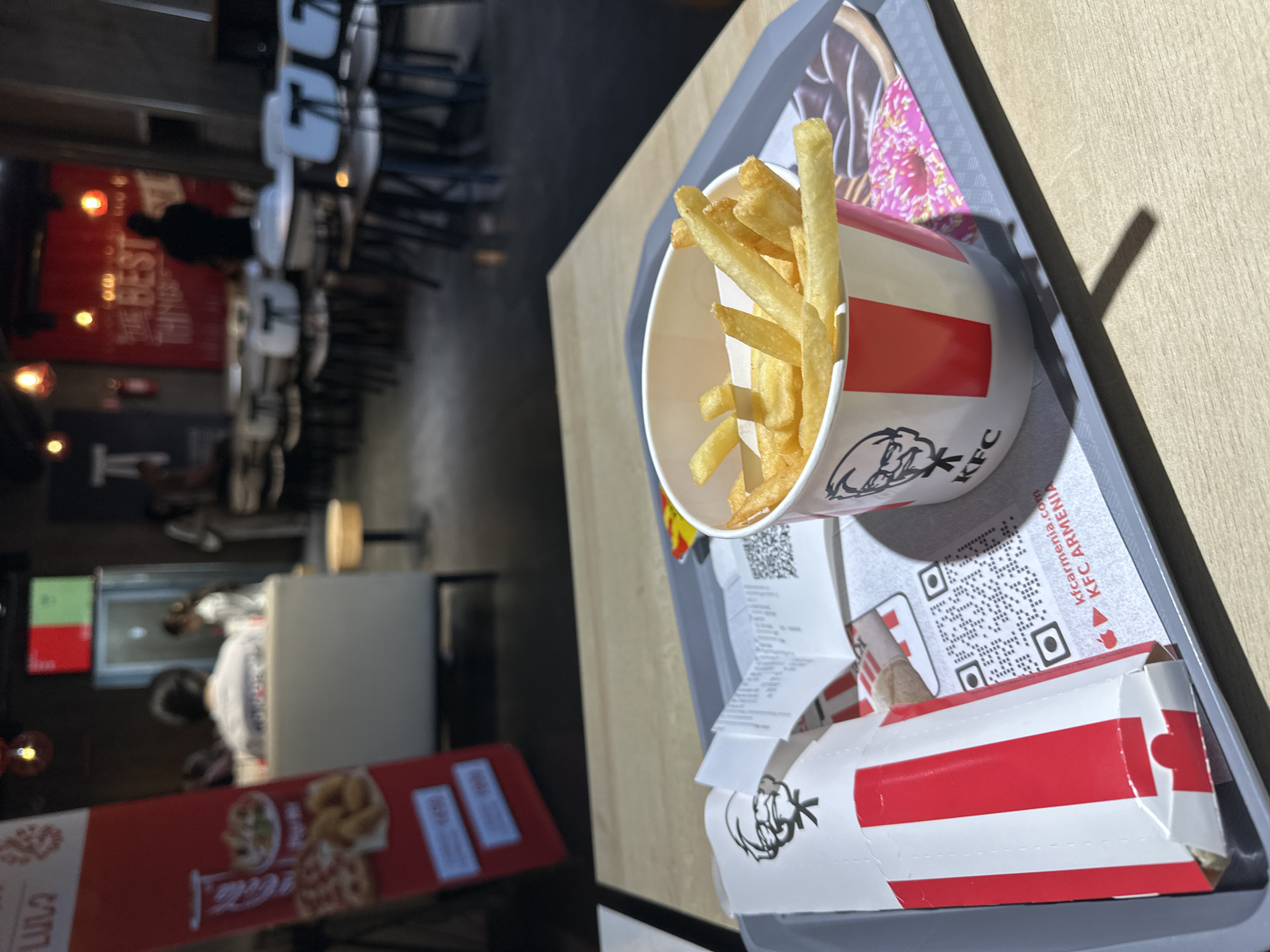 This is a close-up shot inside a KFC restaurant in Yerevan, Armenia. A tray on a wooden table holds a cup of french fries, a wrapped food item, and a receipt. The cup and wrapper feature the KFC logo. The receipt includes the text "KFC ARMENIA" and the website "kfcarmenia.com". In the blurry background, there are tables and chairs, and a menu board. People are visible inside the restaurant.
