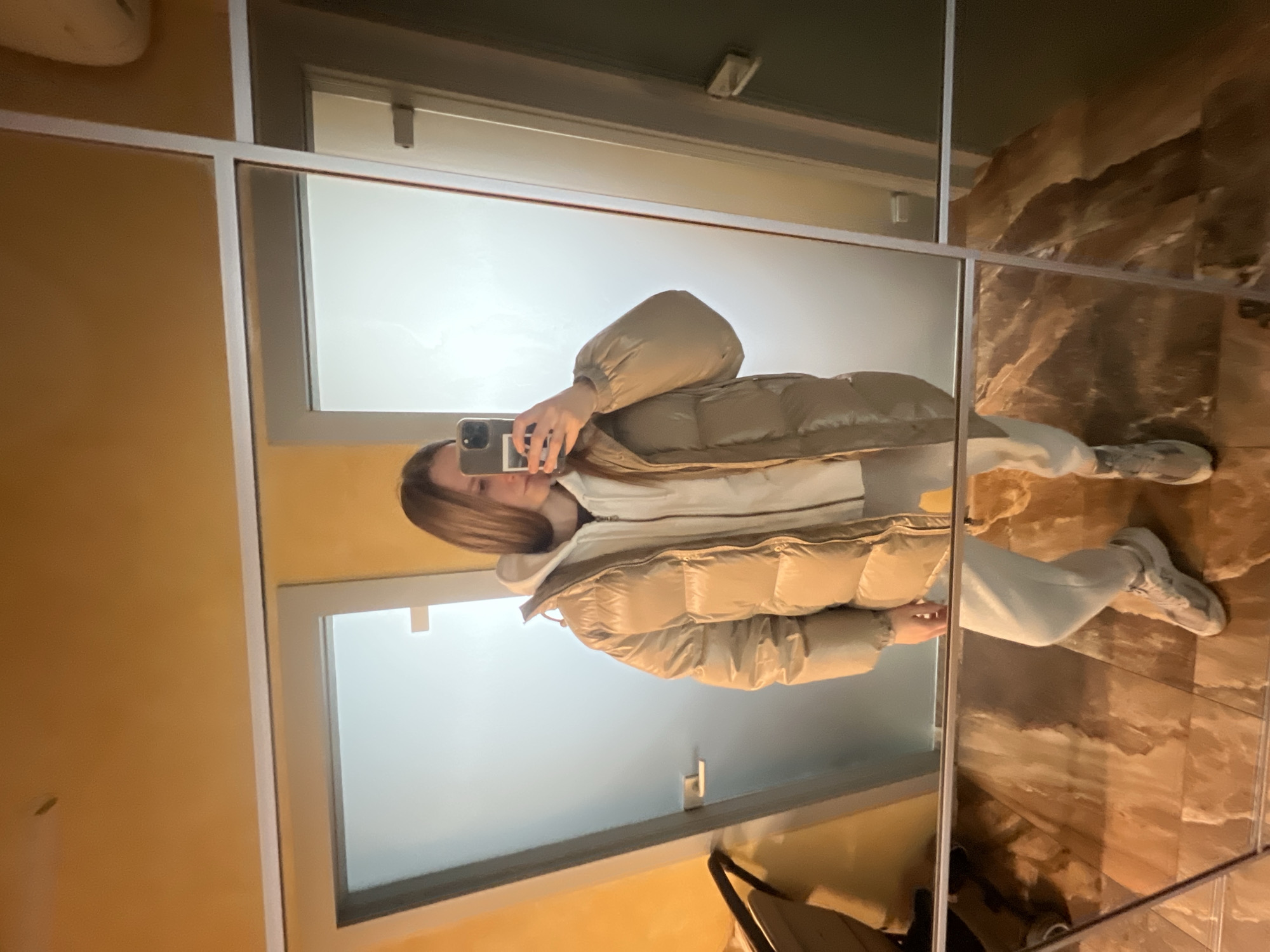 A young person is taking a selfie in a room with a triple mirror. The walls are painted yellow in some sections and have a brown marble finish in others. There is a door reflected in the mirror with a white doorframe. The subject is wearing a beige puffer jacket, a white hooded sweatshirt, and white sweatpants. They are holding a phone in their hand, presumably to take the photograph. The photo was taken in Khimki, Russia.