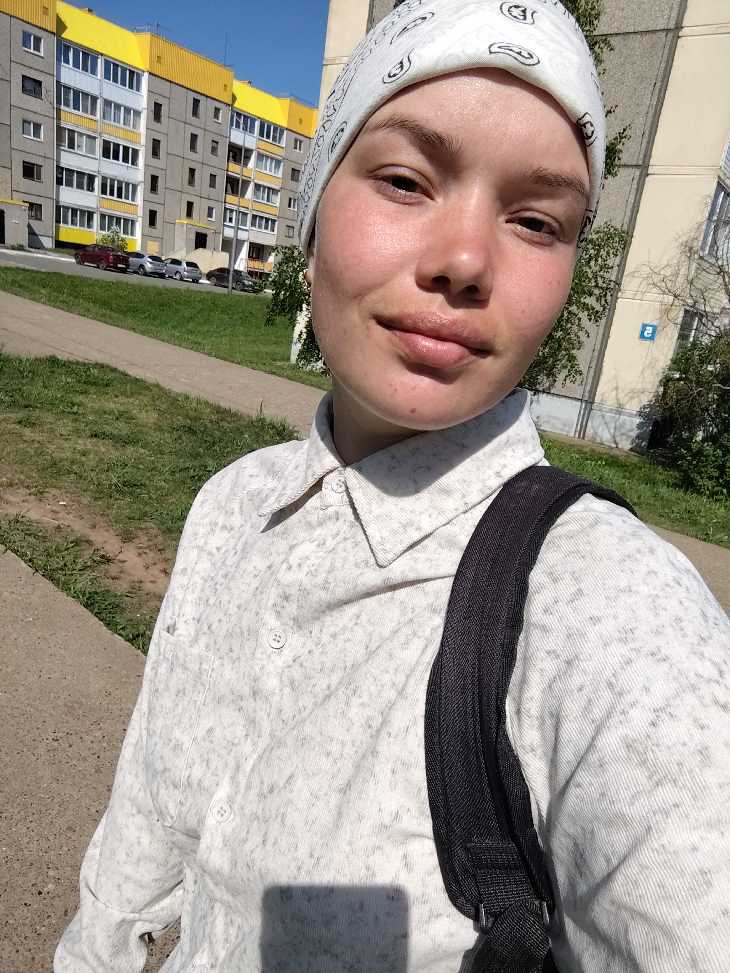 The photograph shows a young woman, likely in her late teens or early twenties, wearing a white headscarf and a light-colored patterned shirt with a collar. She has a dark-colored backpack slung over her shoulder. Her facial features are prominent, and her skin appears to be slightly flushed.
The background includes an apartment building with a distinctive yellow roof, suggesting a Soviet-era construction style. The presence of grass, a sidewalk, and parked cars implies an urban environment. A road with parked cars is on the other side of a green lawn. Based on visual clues and geo-location capabilities, it can be inferred that the photo was taken in the city of Марковский, Russia.