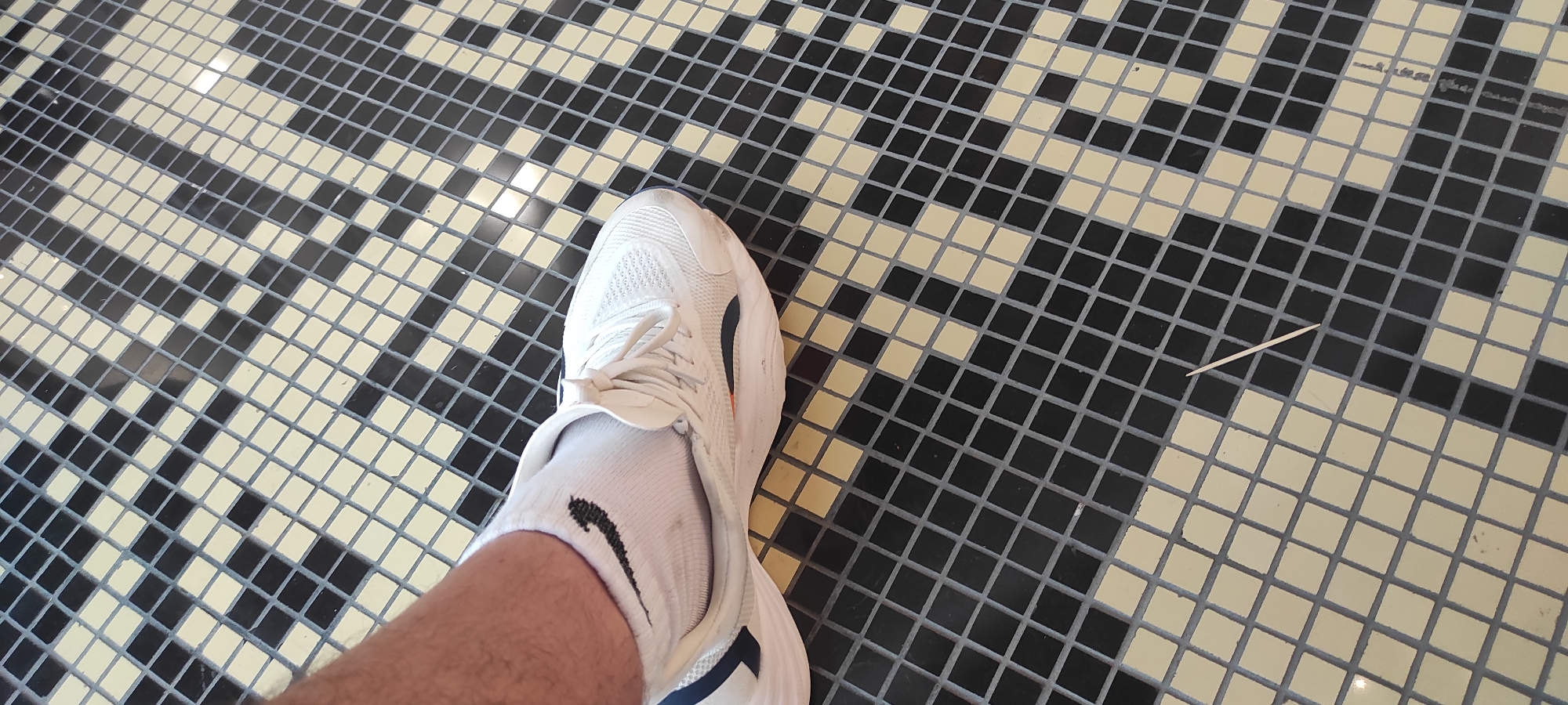 A first-person perspective shot shows a person's left foot, wearing a white athletic shoe and white sock with a black Nike logo, resting on a tiled floor. The floor is covered in small, square tiles arranged in a diagonal pattern with alternating black and pale yellow colors. The tiles form a geometric design. A thin, light-colored toothpick lies on the floor in the lower right portion of the image. The overall impression is of an indoor or covered outdoor space with a decorative tiled surface. The image does not provide any specific geographical context, such as clues to its location in Moscow, Russia.