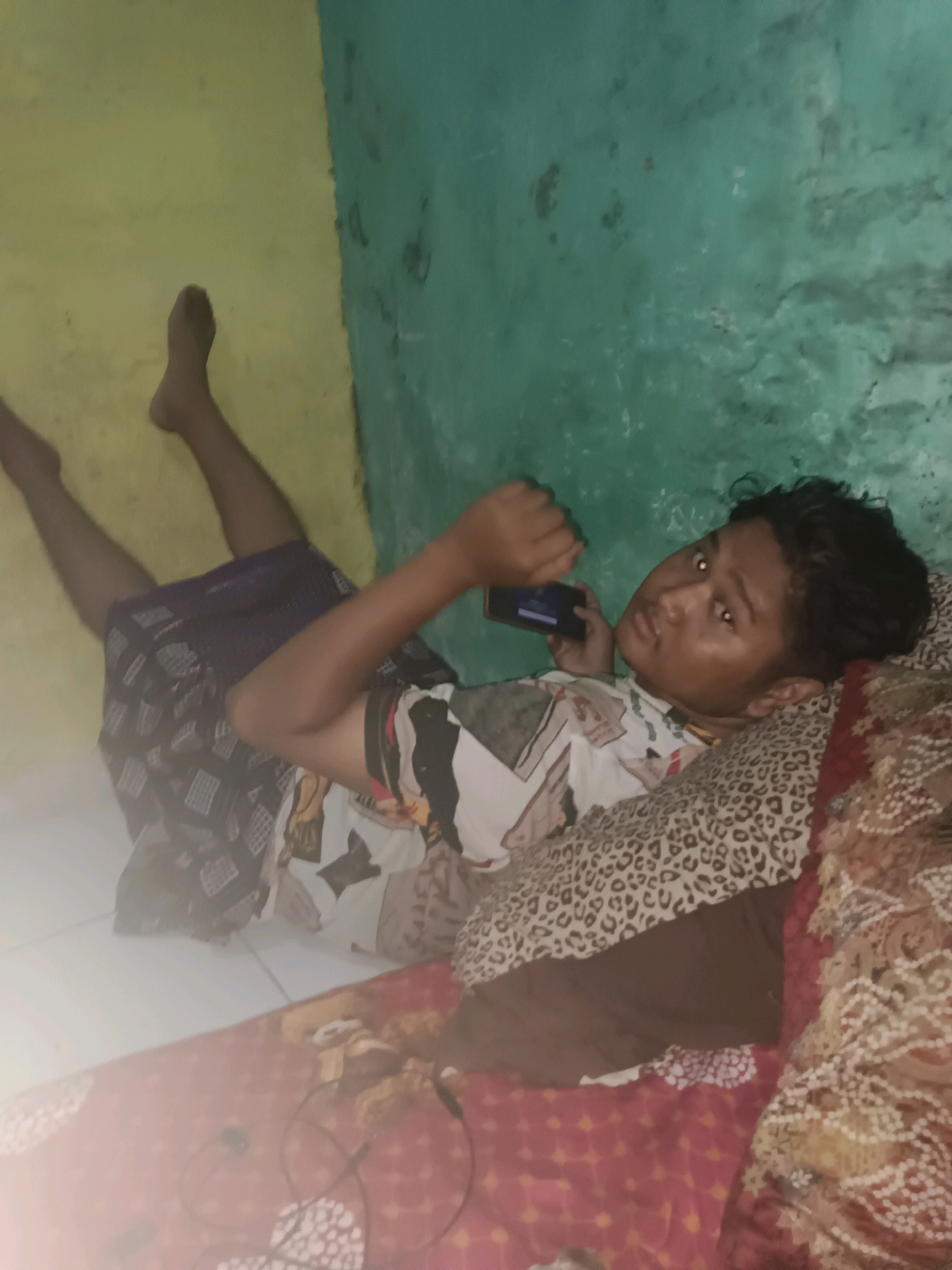 A young male is lying on his back on a bed with a patterned blanket. He is holding a smartphone in his right hand and looking towards the camera. He is wearing a patterned t-shirt and a sarong. The room has green and yellow walls. There are several pillows on the bed. The floor is tiled. The image appears to be taken indoors in the city of Palimanan, Indonesia.