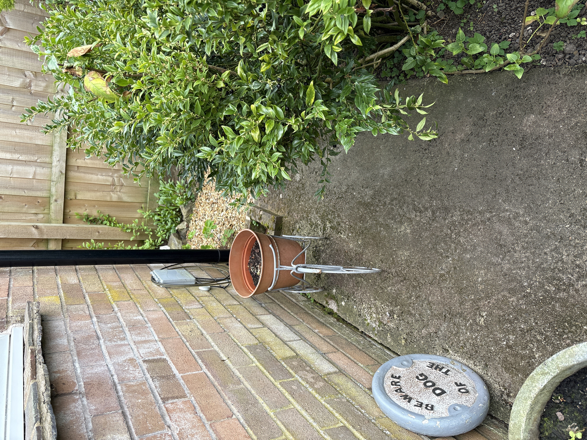 The image depicts an outdoor scene in Longden, United Kingdom. A brick wall runs along the left side of the frame. Adjacent to the wall is a paved area. A brown terracotta pot sits on a metal stand on the pavement. Next to it is a round stone plaque with the inscription "BEWARE OF THE DOG". To the right of the pavement is a green bush. A wooden fence borders the area in the background. A black pipe runs along the top of the brick wall. A cable runs near the wall next to the pipe.