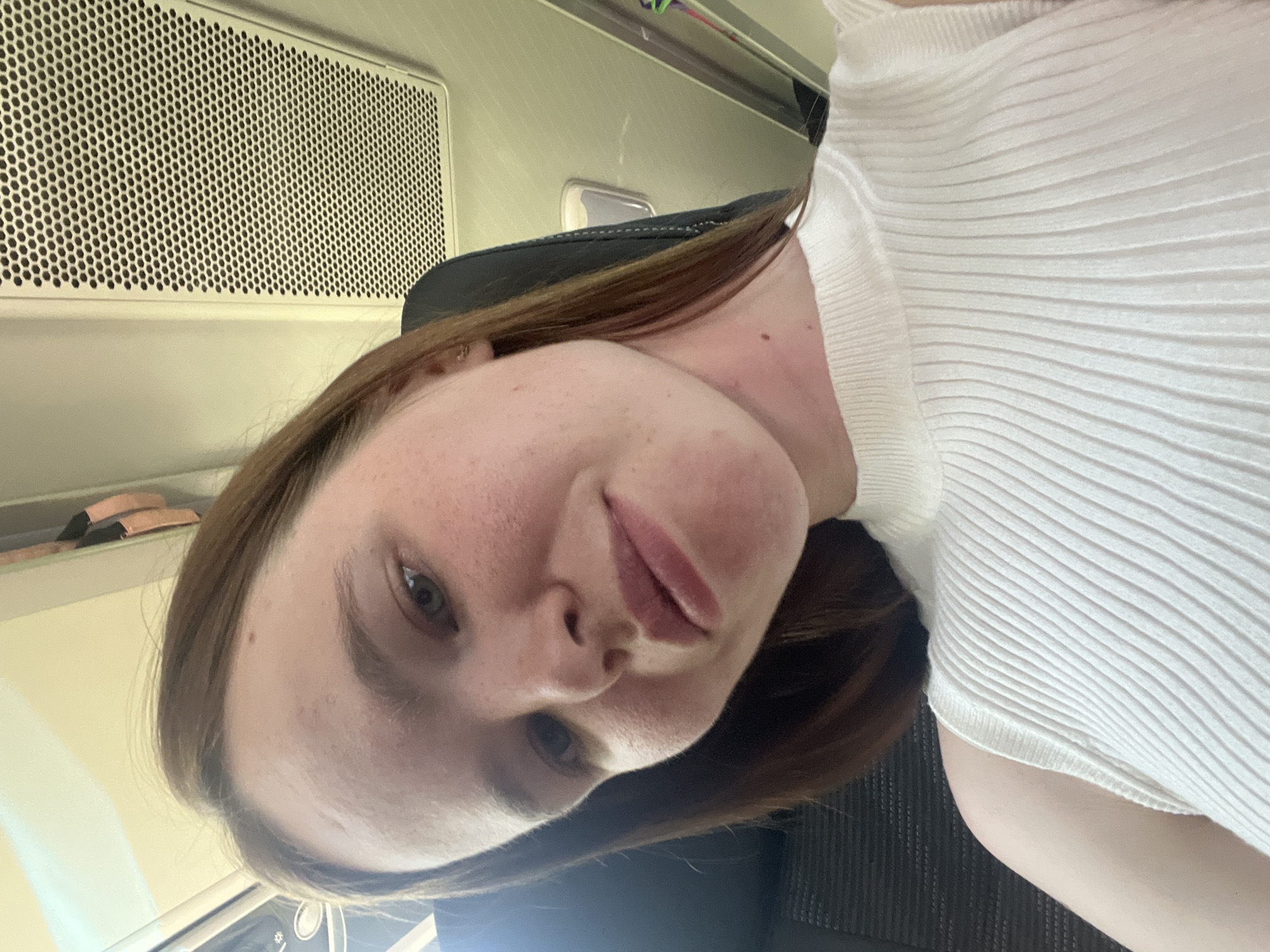 A selfie-style, close-up photograph captures a fair-skinned woman with light brown hair, wearing a white ribbed top. The background suggests she is likely seated on an airplane, with a ventilation panel visible above and behind her head. The scene appears to be indoors and well-lit, but specific location details in Alnashi, Russia are not discernible.