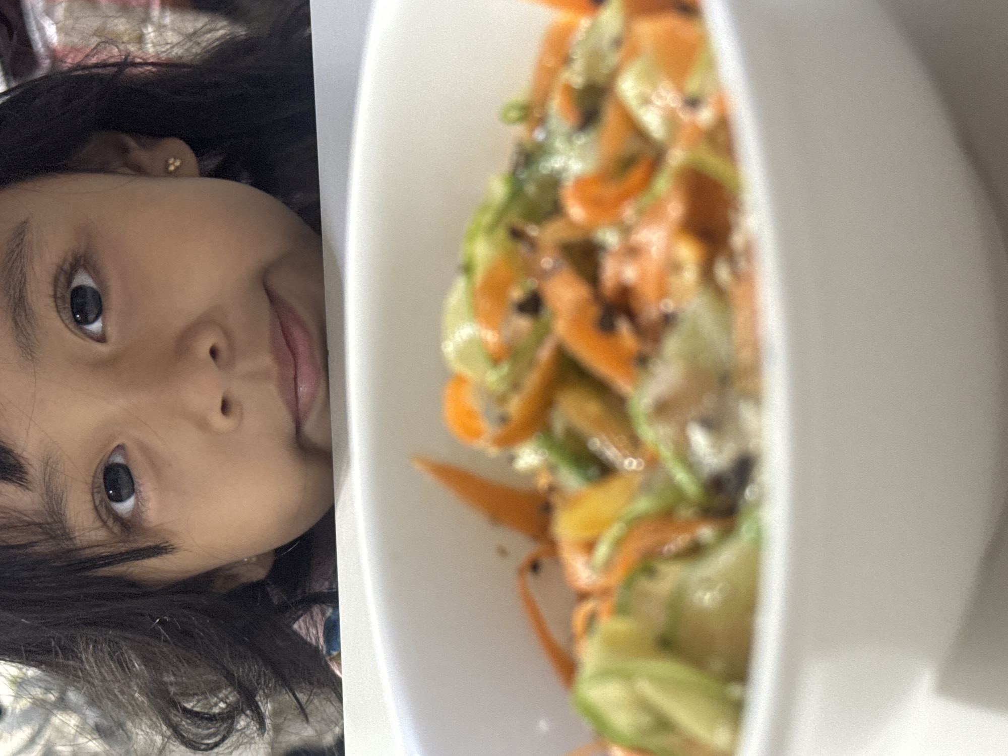 A young child, appearing to be female with dark, wavy hair and medium skin tone, is positioned at the top of the frame, peering over a white surface. Her dark brown eyes are directed towards the viewer, and she exhibits a subtle, closed-mouth smile. A small, gold-colored stud earring, composed of three tiny spheres, is visible in her left earlobe. Her upper body and the background behind her are heavily blurred, with only hints of a multi-colored garment visible on her chest.
In the foreground, occupying the lower half of the image, is a white ceramic bowl containing a dish of mixed vegetables. The food is out of focus, appearing as soft blurs of vibrant orange and green, consistent with julienned carrots and sliced cucumbers or zucchini. Small dark specks, possibly sesame seeds or spices, are scattered throughout the dish. The bowl rests on a flat, white surface, which also serves as a barrier between the food and the child. The immediate background behind the child is indistinct and heavily blurred, suggesting an indoor setting without specific identifiable features. The scene is located in Davao City, Philippines.