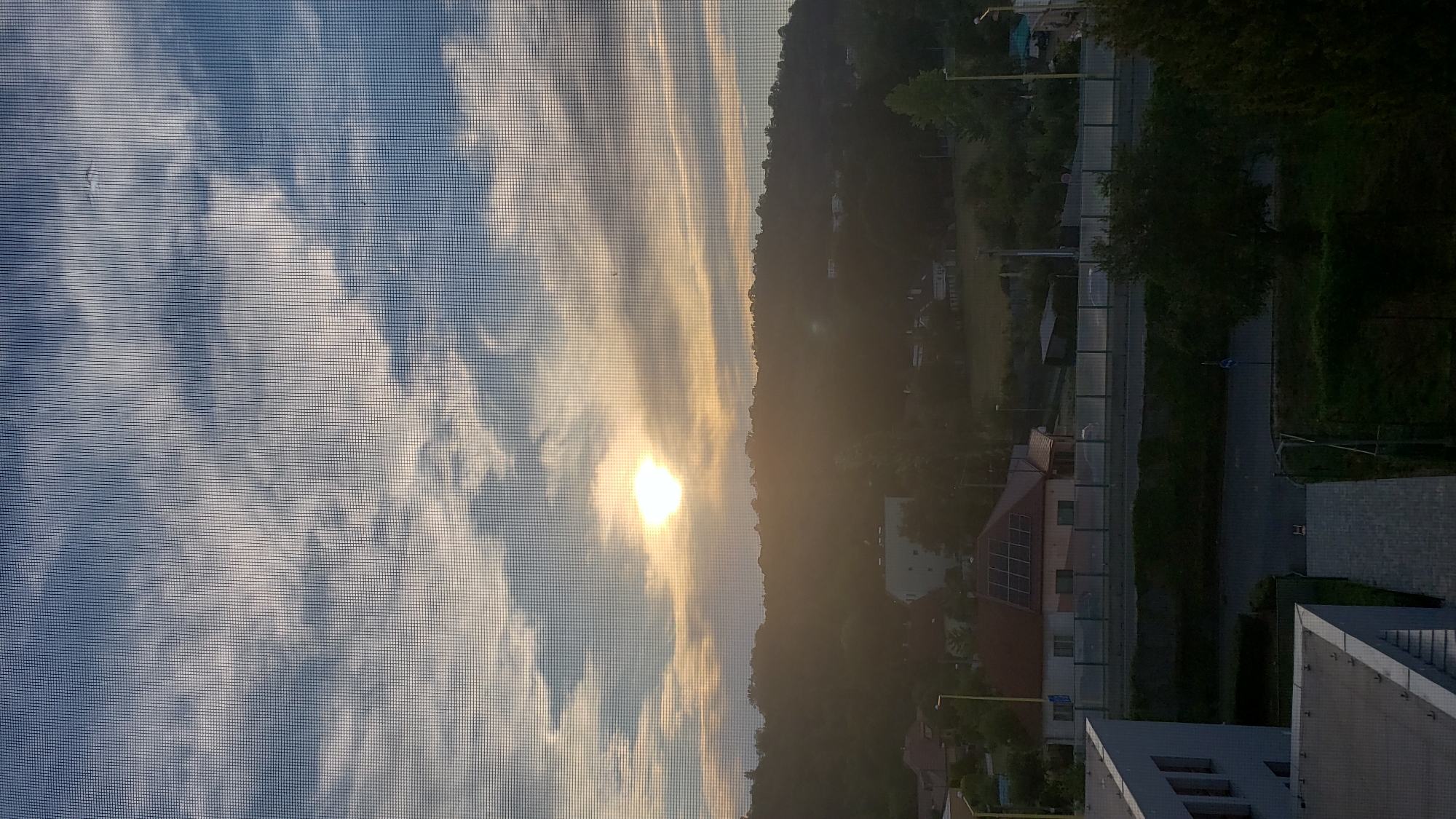 This image captures a sunrise viewed through a window screen in Prešov, Slovakia. The foreground shows parts of modern buildings, including a white structure with visible windows on the left. Below these buildings, a road is visible with a low barrier. Further into the scene, a residential area with houses, one featuring solar panels on its roof, is nestled against a tree-covered hill. The sky is a prominent feature, with the sun appearing as a bright orb peeking through scattered clouds, casting a warm, hazy light over the landscape. The overall impression is of an urban environment at the start of a new day.