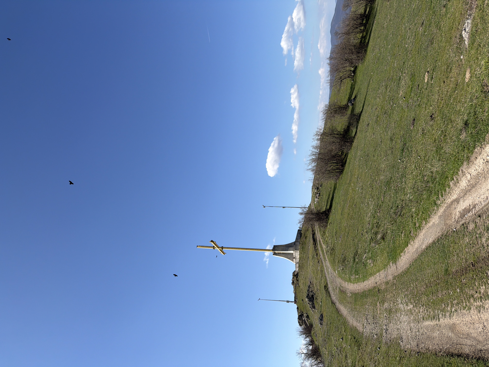The image shows a large, golden cross monument atop a grassy hill in Bata, Bulgaria. A winding dirt path leads up the hill. The sky is blue with some scattered white clouds. Two birds are flying in the sky. There are also two tall, thin, metal poles standing on the hill. Bare trees dot the landscape.
