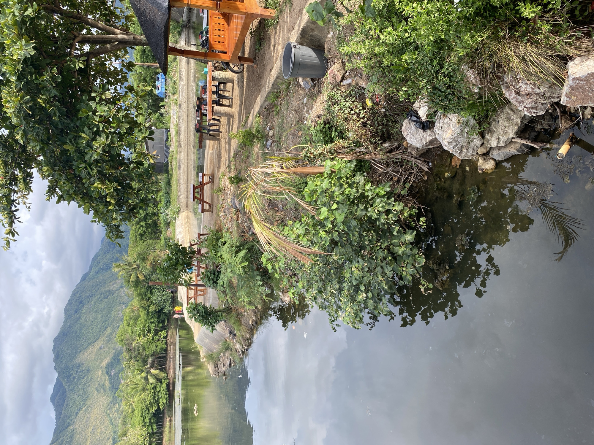 The image depicts a tranquil lakeside scene in Aneuk Paya, Indonesia. A calm body of water reflects the overcast sky and surrounding lush greenery. On the shore, several picnic tables and benches are arranged, suggesting a recreational area. A charming wooden gazebo with a dark roof stands prominently, offering shade. In the distance, rolling hills and mountains form a majestic backdrop. A few people are visible near the water's edge, adding a touch of human presence to the serene landscape. The overall atmosphere is peaceful and natural, highlighting the beauty of the Indonesian countryside.