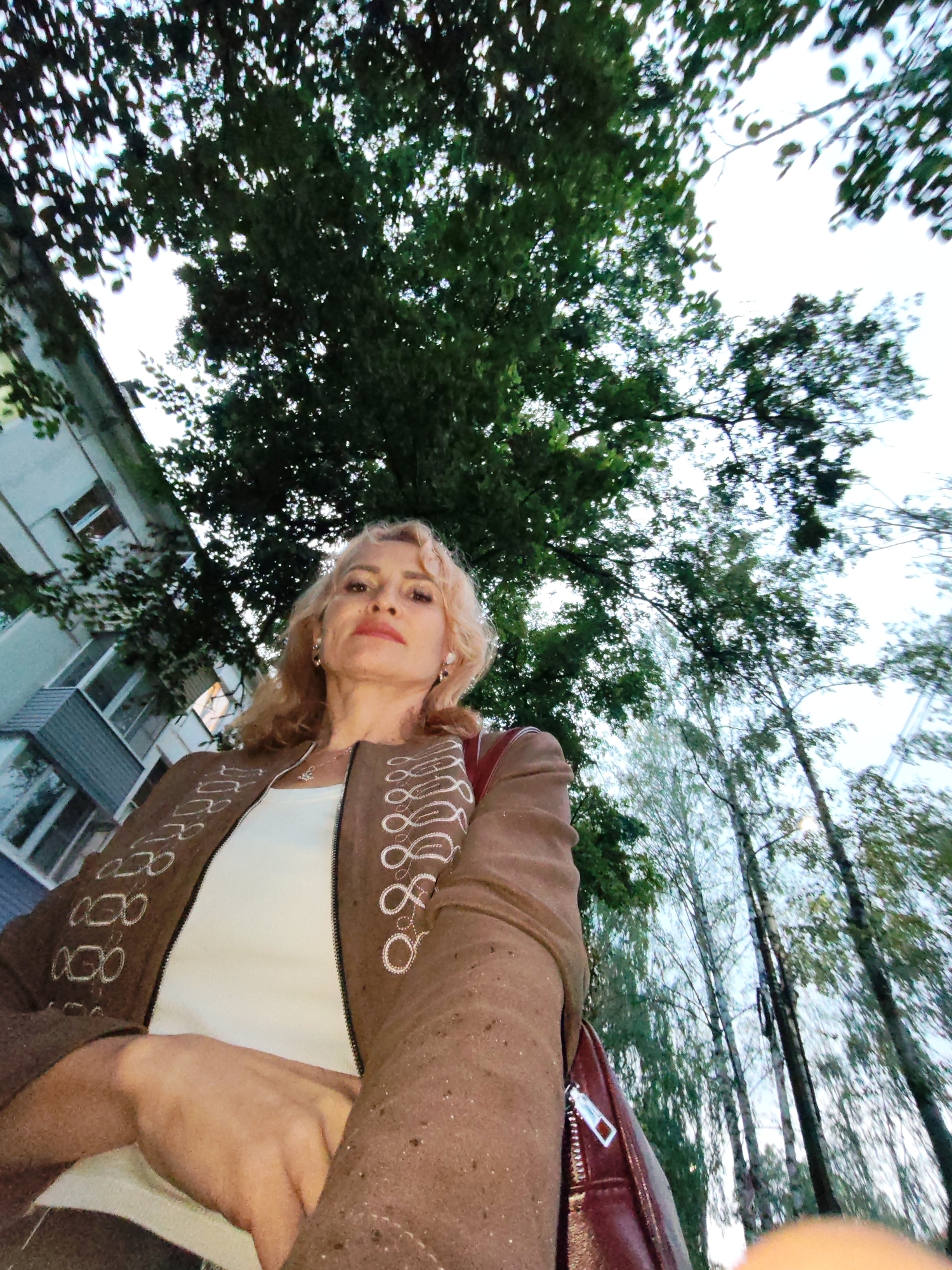 A woman with blonde hair is standing outside in a low-angle shot, looking at the camera. She is wearing a brown jacket with white embroidery, a white top, and a necklace. The background shows a building on the left and trees with green leaves on the right. The lighting suggests it is daytime. The location is the city of Chaikovsky, Russia, as indicated by the urban environment with buildings and trees.