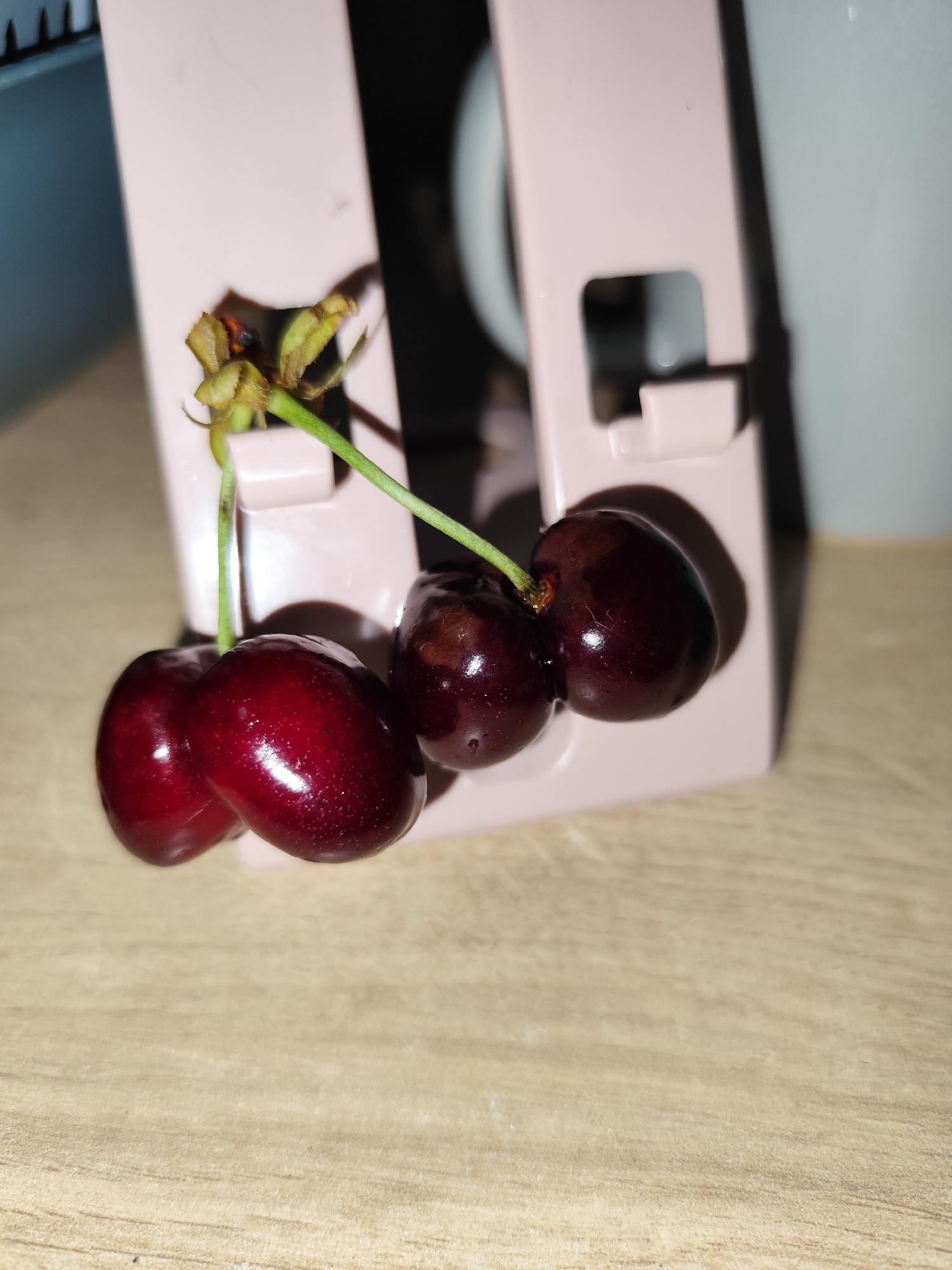A still-life image shows three dark red cherries connected by green stems, one pair attached to a dried, brownish cluster of stems and leaves. They sit in front of a pink plastic stand on a light brown, wood-grain surface. The background includes parts of what appear to be a white mug and a blue container. The location of the image is in the city of Unknown, Unknown.