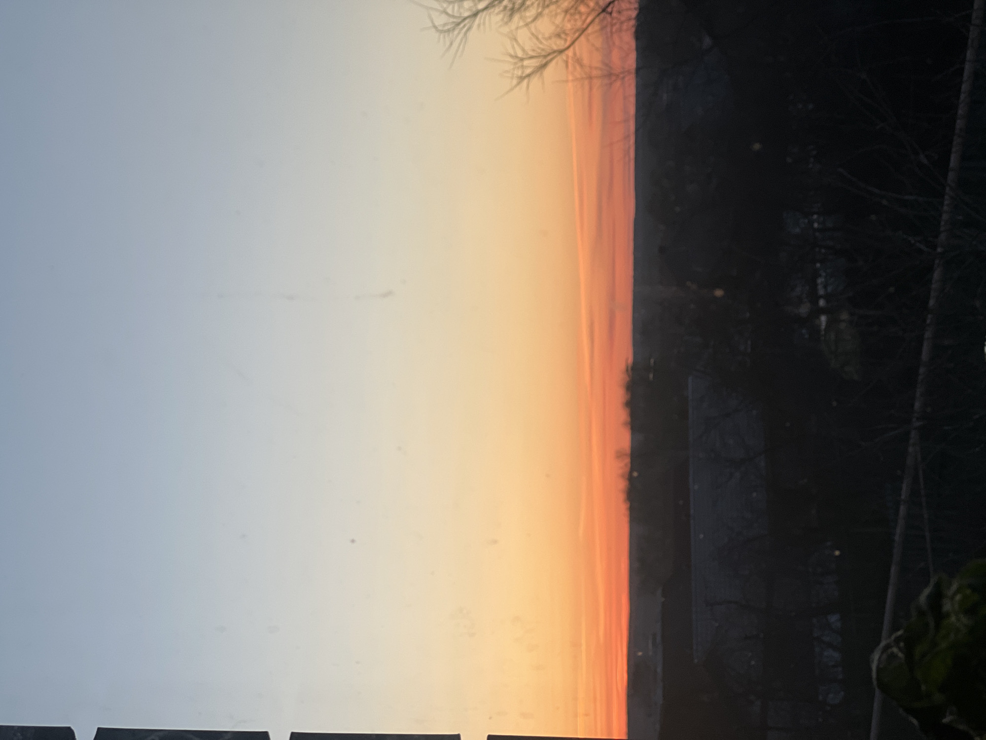 The image shows a sunrise or sunset scene. The sky transitions from a light blue to orange and red hues near the horizon. A dark silhouette of trees and possibly buildings is visible on the right side of the image. There is no evidence to suggest that this is the city of Votkinsk, Russia. It is unclear whether people are present.