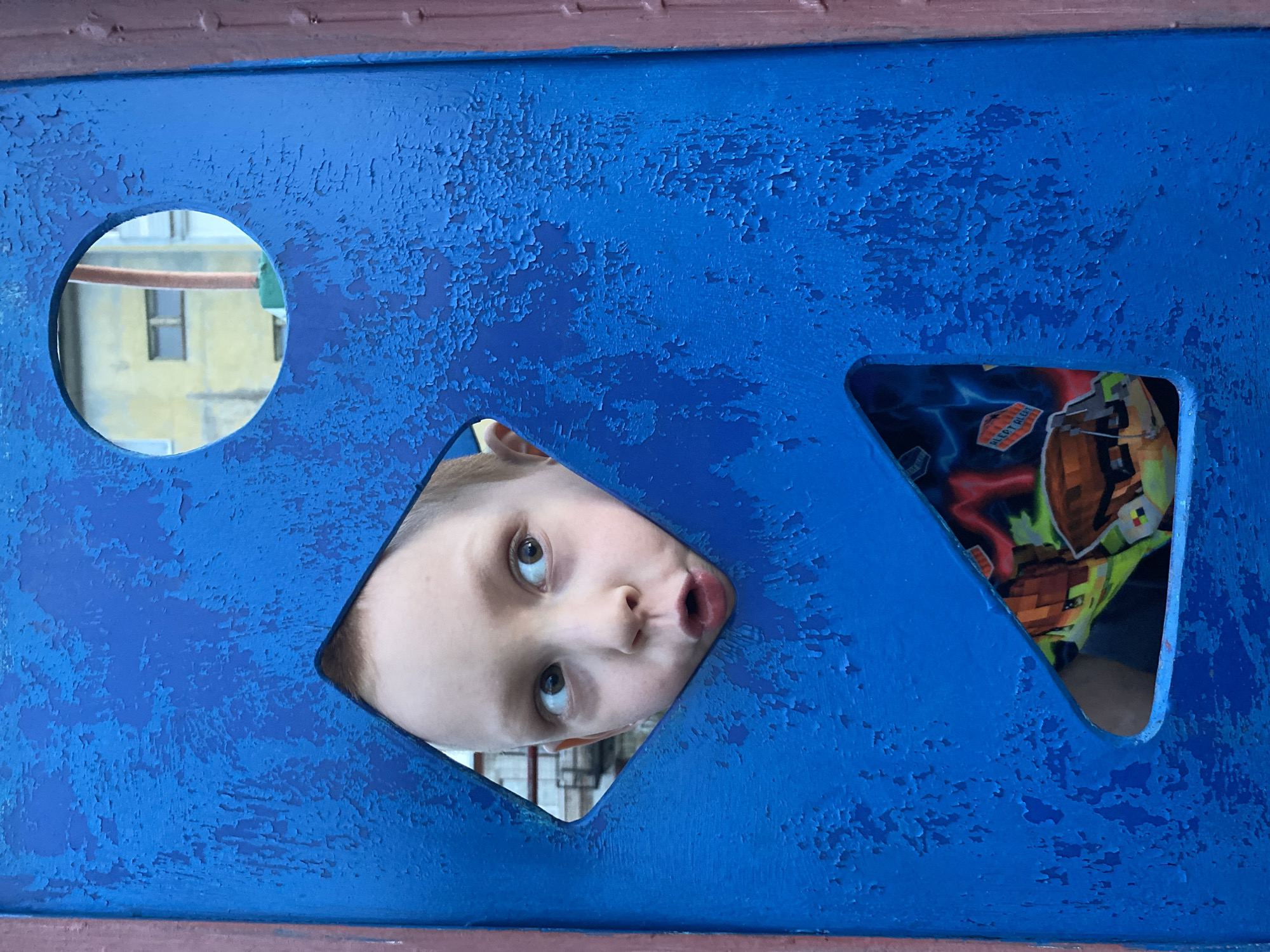A child looks through a square-shaped hole in a blue structure on a playground. The structure also has a round hole and a triangular hole, through which glimpses of the surrounding environment and the child's clothing can be seen. The blue paint on the structure is peeling, indicating its age and exposure to the elements. A building is visible through the round hole. Based on visual data, the location may be Noviy, Russia.