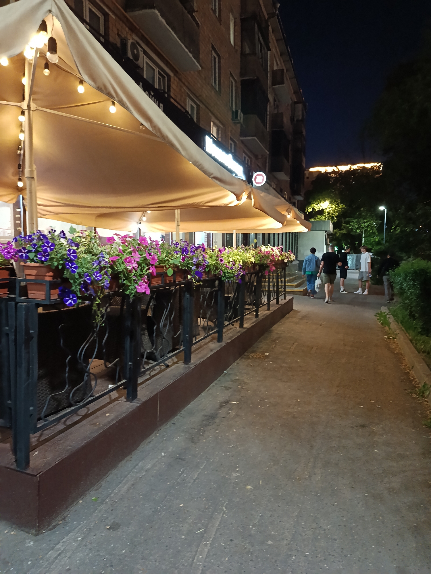 A nighttime shot shows a sidewalk adjacent to a building with a lit outdoor cafe or restaurant in Moscow, Russia. The building is multi-storied and appears to be residential or mixed-use, with balconies and windows visible. An awning covers the cafe's outdoor seating area, illuminated by strings of lights. Flower boxes are attached to a wrought-iron fence that separates the sidewalk from the cafe's seating. A group of people are walking down the sidewalk away from the camera; they are dressed in casual attire. A grassy area borders the sidewalk on the right, with trees and a street light further in the background. The sky is dark, suggesting the photograph was taken in the evening.