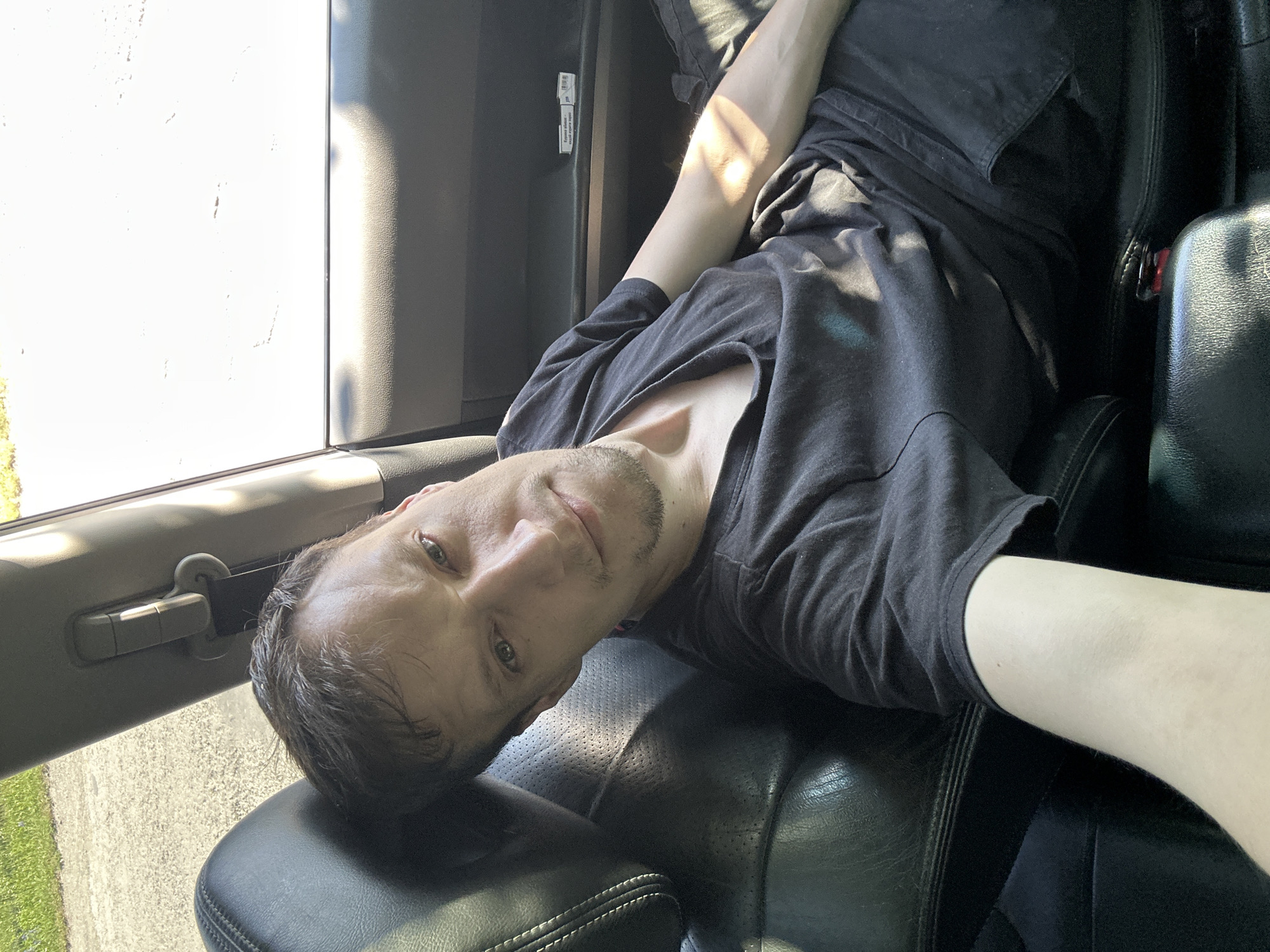 The image shows a man sitting in the passenger seat of a car, likely on a sunny day. He is wearing a black t-shirt and dark pants. His left arm is extended, holding what appears to be a phone, suggesting he is taking a selfie. The interior of the car is visible, including black leather seats and part of the dashboard. Sunlight streams into the car, creating highlights on the man's arm and clothing. Outside the car, a blurred background shows a paved surface, possibly a road or parking lot, with green grass and trees visible on the left side of the image. The location is specified as Shatava, Ukraine.