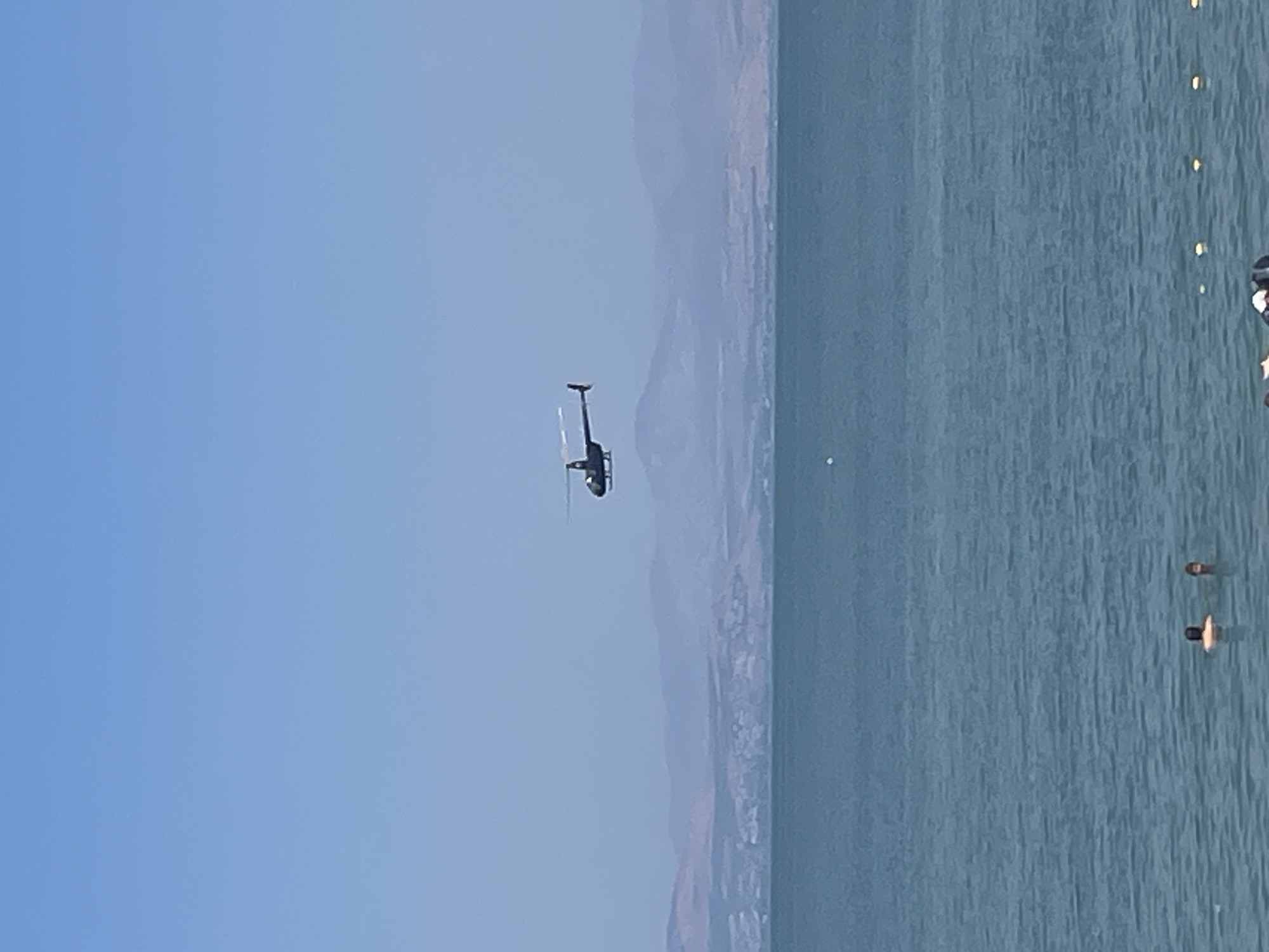 A helicopter is flying over the Sea of Galilee, a large freshwater lake in Emek HaYarden Regional Council, Israel. The water is a calm, deep blue, with gentle ripples on its surface. In the foreground, two people are visible swimming in the lake. In the background, rolling hills and mountains can be seen, shrouded in a light haze under a clear, pale blue sky. The helicopter is a dark color and appears to be a smaller model, with its rotor blades extended.