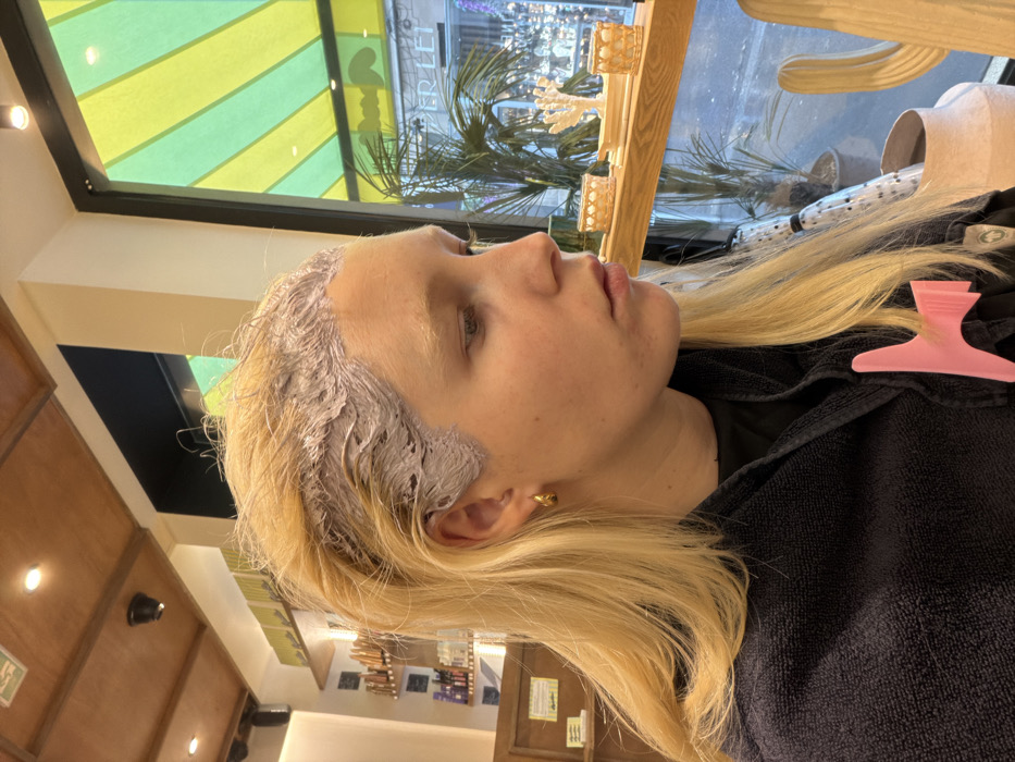 A fair-skinned individual, likely female, with long blonde hair, is seated in a hair salon, captured in a profile view facing right. A light purple or grey chemical substance, consistent with hair dye or toner, is applied to the roots of her hair, particularly visible along the hairline and scalp. She wears a dark blue or black protective salon cape, to which a pink, claw-shaped hair clip is affixed on her right chest area. A small, gold-colored hoop earring is present in her visible left ear. Her gaze is directed slightly downward.
The interior environment features a wooden paneled ceiling with recessed lighting. In the blurred background on the left, shelves stocked with various hair products are visible. To the right, a large window provides a view of the exterior. Outside the window, a distinctive awning with alternating vertical stripes of bright green and yellow is prominent. Below the awning, partially visible text reads "PER LEI". The windowsill hosts several decorative objects, including a white, branched coral-like sculpture, two small woven basket-like containers, and a light-colored, possibly ceramic, cactus sculpture. Beyond the window and decorations, a potted plant with large leaves and blurred elements of a city street are visible. The establishment is identified as Per Lei Coiffure - Esthétique, located at 8 Rue de Caumartin, 75009 Paris, France.