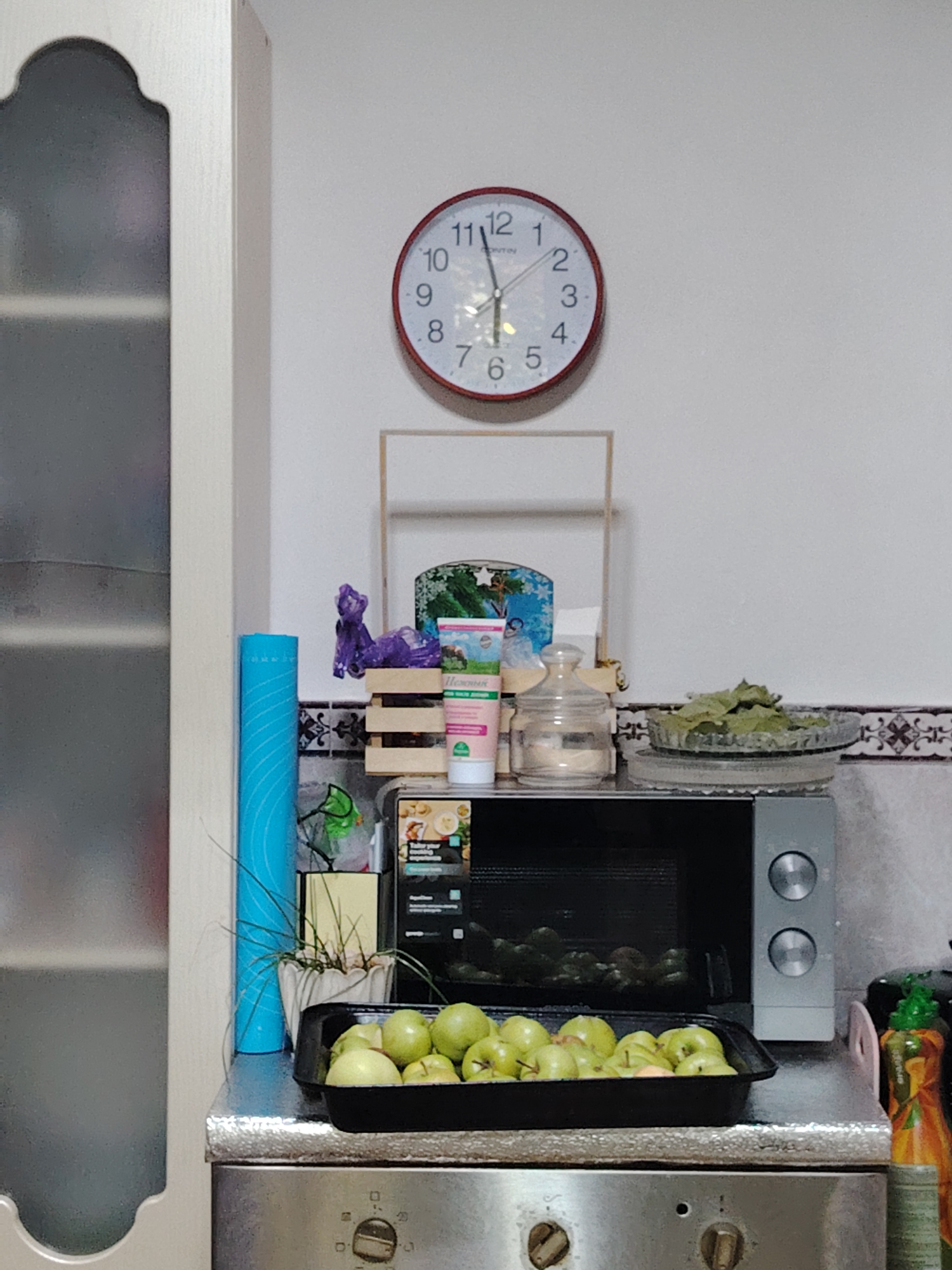The image shows a cluttered kitchen counter with a microwave, a bowl of green apples, and various other items. A round clock with a red frame hangs on the wall above the counter. To the left, a portion of a tall, white cabinet with a decorative curved top is visible. A bright blue cylindrical object, possibly a mat or a roll of material, stands upright next to the cabinet. A small potted plant with green leaves is situated behind the blue cylinder. On the counter, a black tray is filled with numerous green apples. Behind the apples, there is a microwave oven. To the right of the microwave, a glass jar with a lid is present, next to a small wooden crate holding some items. A decorative item with a picture of a snowy landscape and a winter scene is leaning against the crate. A few tubes or bottles are also on the counter. The background wall appears to be white with a decorative tile border near the counter. The overall impression is a domestic kitchen setting.