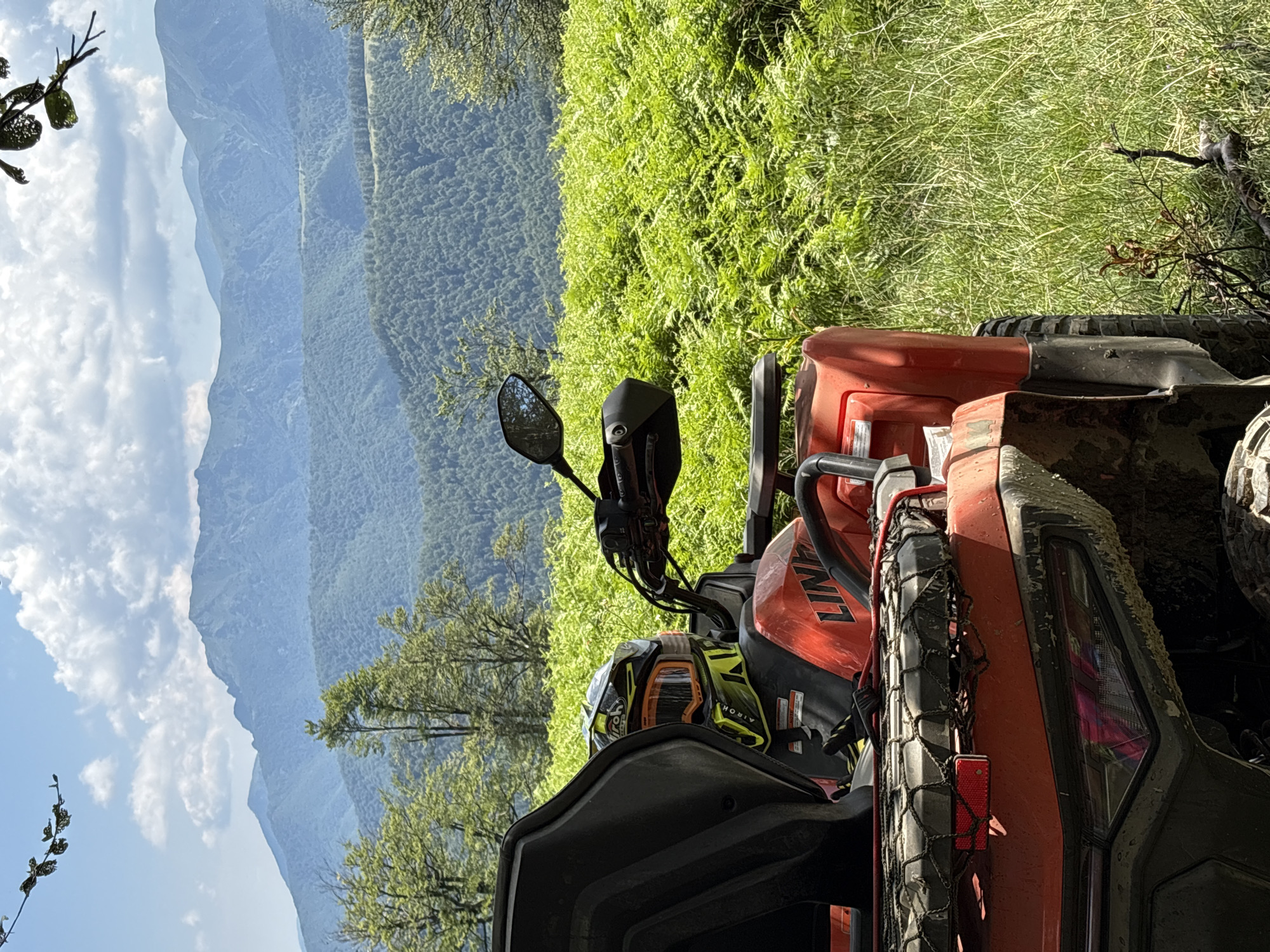 In Apriltsi, Bulgaria, an orange ATV is parked on a grassy slope with a dense forest of trees covering rolling hills in the background. The ATV has a black seat with a yellow and black helmet and goggles resting on it. The handlebars are visible, as well as a side mirror reflecting the sky and trees. The ATV is positioned to the right of the frame, with its rear and side visible. The terrain is covered in vibrant green ferns and tall grass. The sky is blue with scattered white clouds. In the far distance, a mountain range is visible, with layers of forested slopes and rocky peaks. The scene appears to be a natural, outdoor environment, possibly a hiking or off-roading trail.