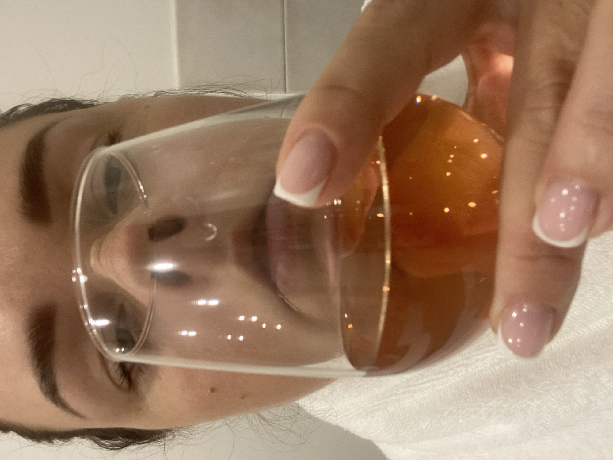 A fair-skinned person is shown in a close-up shot. The person's face is visible behind a clear glass, which contains a transparent amber-colored liquid. The person is holding the glass with their right hand, and their fingers are adorned with pale pink nails, each with a white tip. The background appears to be a white tiled wall, with a white surface underneath. There is no clear indication of the location being Sochi, Russia from the image.