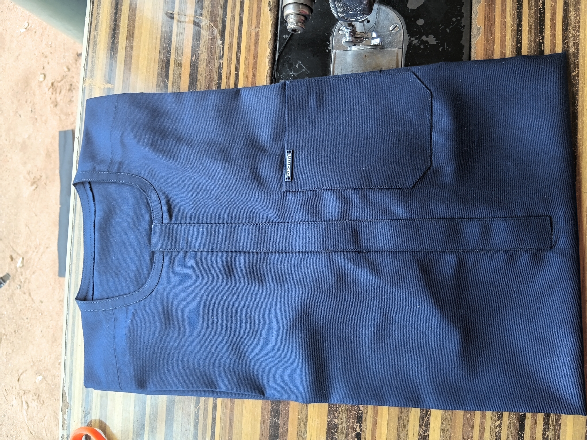 A dark blue long-sleeved shirt is folded flat on a wooden surface. The shirt has a rounded neckline with a small placket down the center front. A single breast pocket is visible on the left side of the shirt, adorned with a silver-colored rectangular metal tag that reads "EXECUTIVE." The wooden surface appears to be a table with a worn, striped pattern. A portion of a black sewing machine is visible on the right side of the image, suggesting the shirt may have been recently made or is being prepared for further alteration. The background is out of focus, but appears to be an outdoor setting with sandy ground. The location is identified as Jalingo, Nigeria.